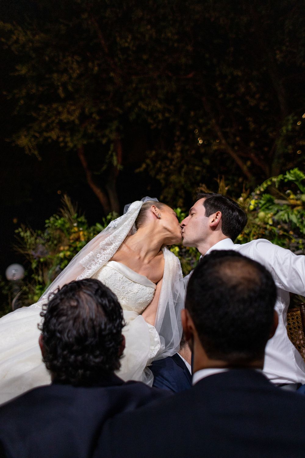 Romantic wedding kiss moment captured from behind guests during an evening outdoor ceremony.