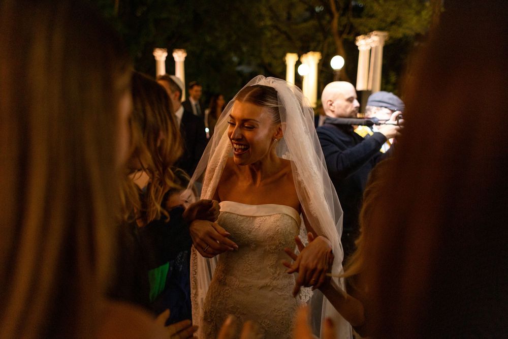 A bride in a strapless white wedding dress and veil shares a candid moment during an evening outdoor celebration.
