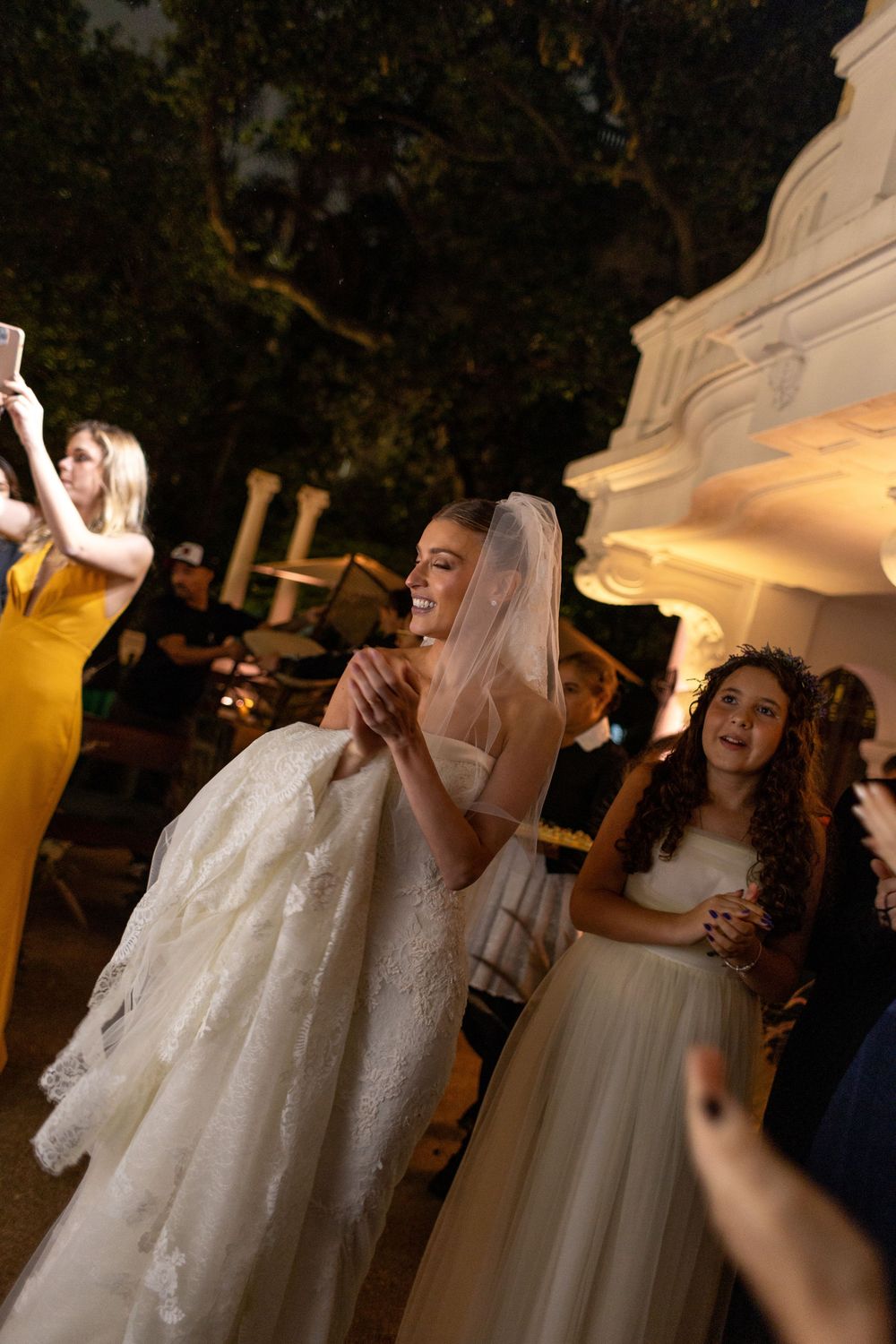 A bride in a white lace dress celebrates joyfully during an outdoor evening reception.