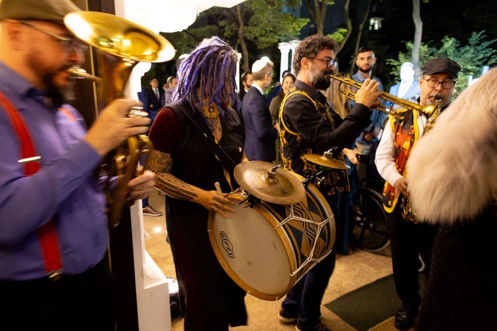 Wedding guests gather around musicians playing brass instruments at an outdoor celebration.