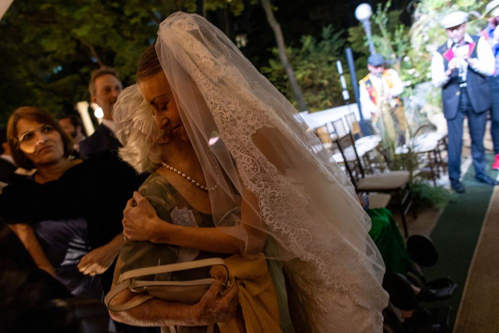 A bride in a flowing veil leans forward during an outdoor evening celebration with guests visible in the background.
