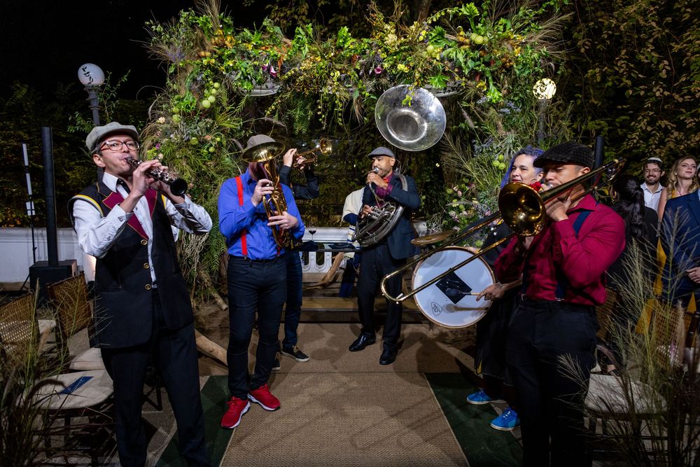 A lively brass band performs outdoors under garden lights and greenery during an evening celebration.
