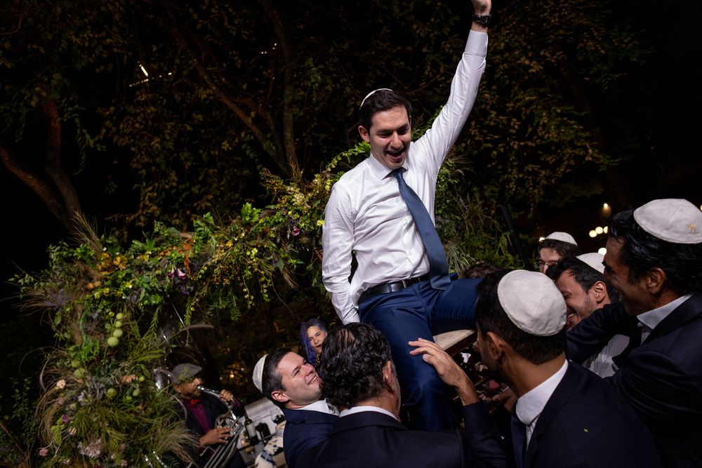 A group dances joyfully during an outdoor Jewish wedding celebration beneath trees and natural greenery.