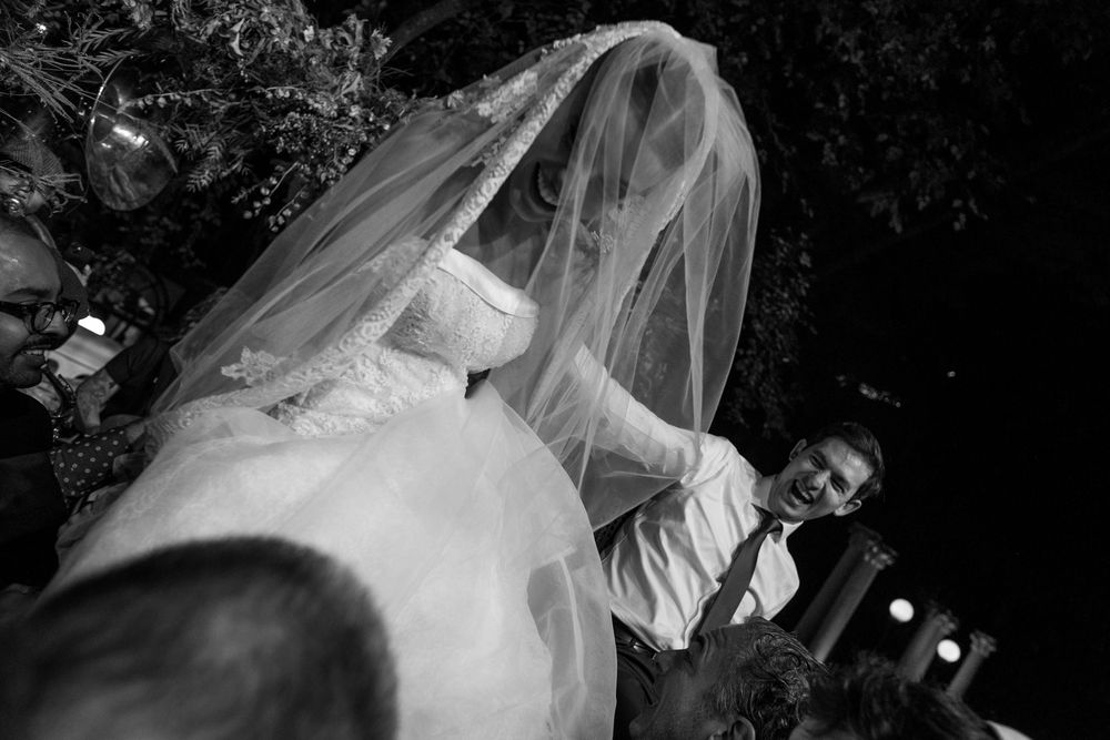 A black and white wedding photo showing a bride in a flowing veil with a figure visible in the background.