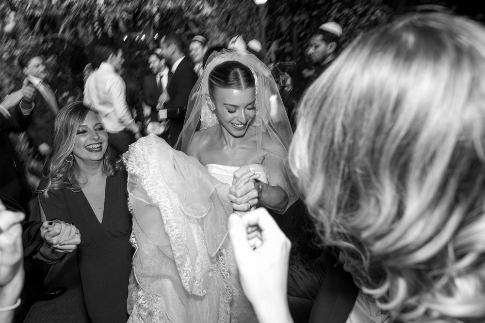 A bride in a white wedding dress laughs while dancing with guests at a reception in black and white photography.