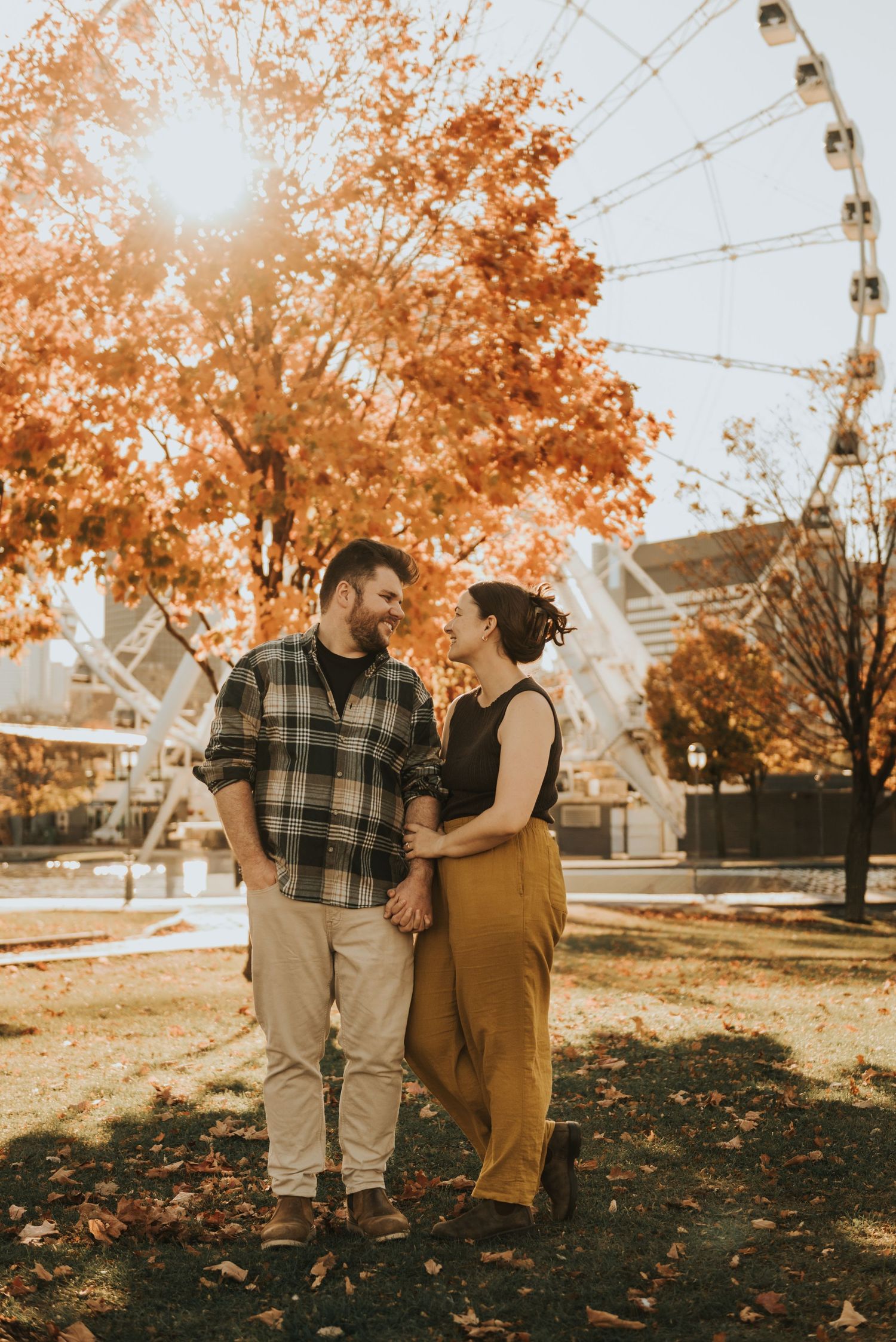 A couple enjoys an autumn day with orange foliage and a ferris wheel in the background at a park.