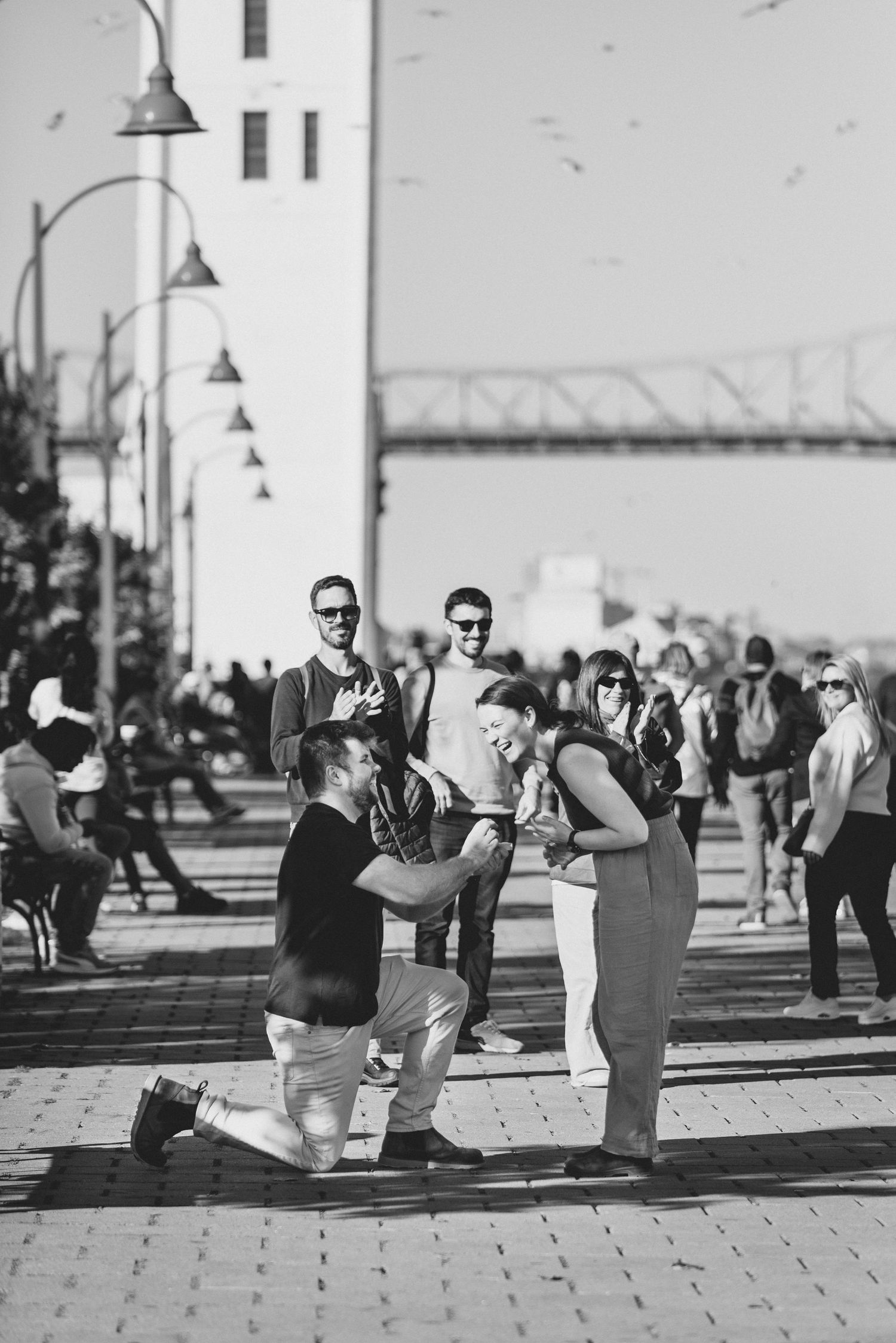 A romantic proposal moment captured in black and white on a brick-paved walkway with lamp posts and a bridge in the background.