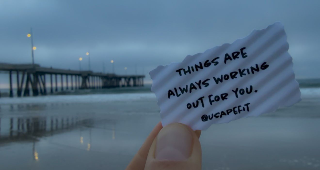 A handwritten note reading 'Things are always working out for you' held up against a moody pier backdrop at dusk.