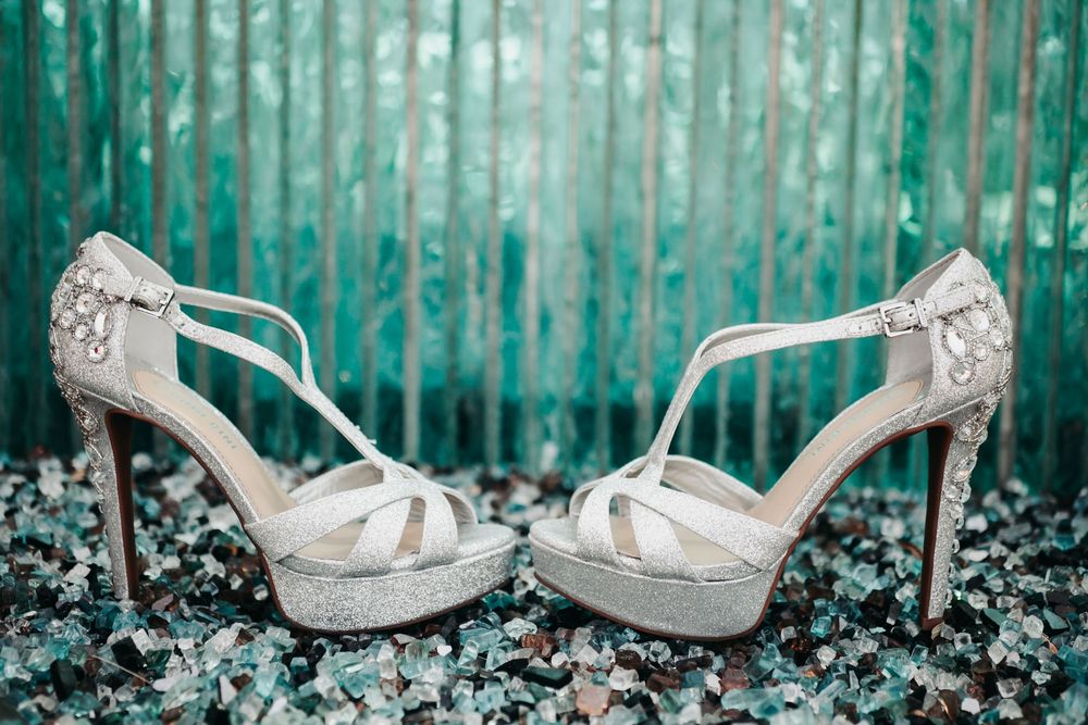 Elegant white lace bridal heels with peep toes and strappy details displayed against a turquoise corrugated background.