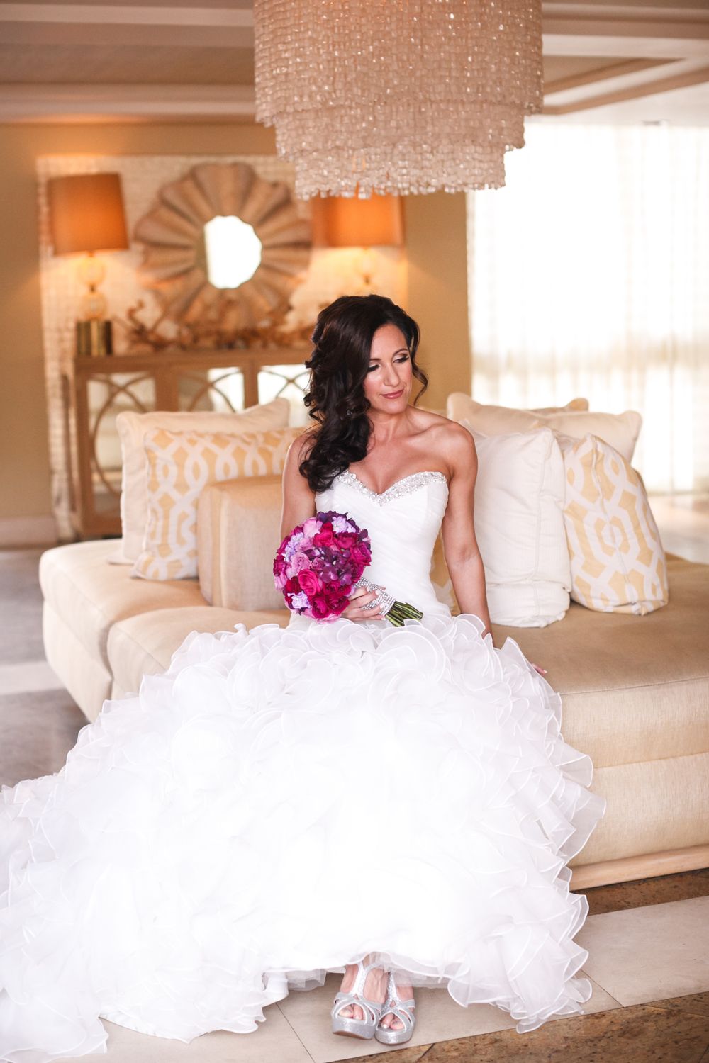 A bride in a voluminous white wedding gown with purple flowers lounges elegantly on a cream-colored sofa.