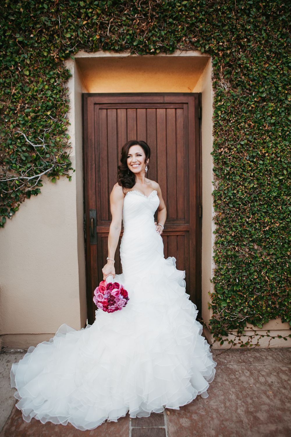 Elegant bride in white ruffled wedding gown poses in front of rustic wooden door with ivy-covered wall backdrop.