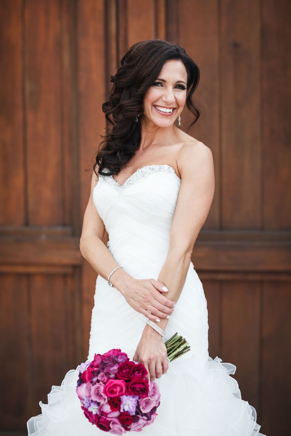 A bride in a white wedding dress holds a vibrant pink and purple bouquet against a rustic wooden backdrop.