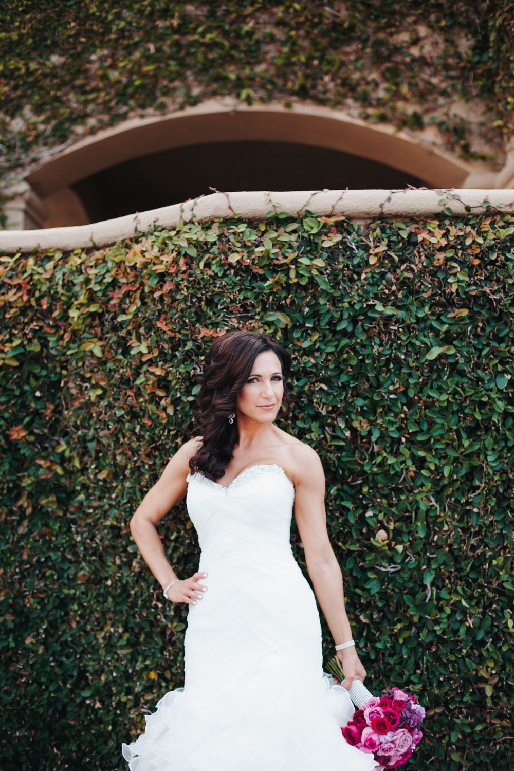 A bride in a white wedding gown poses against a lush green hedge wall while holding a vibrant pink bouquet.