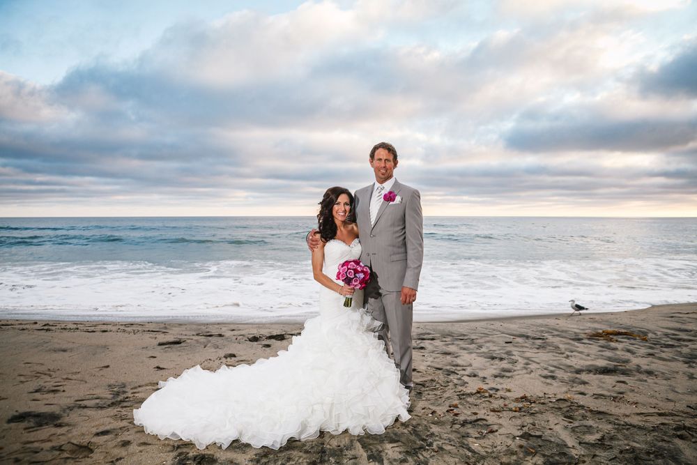 A romantic beach wedding photoshoot with newlyweds posing against the backdrop of a dramatic cloudy sunset over the ocean.