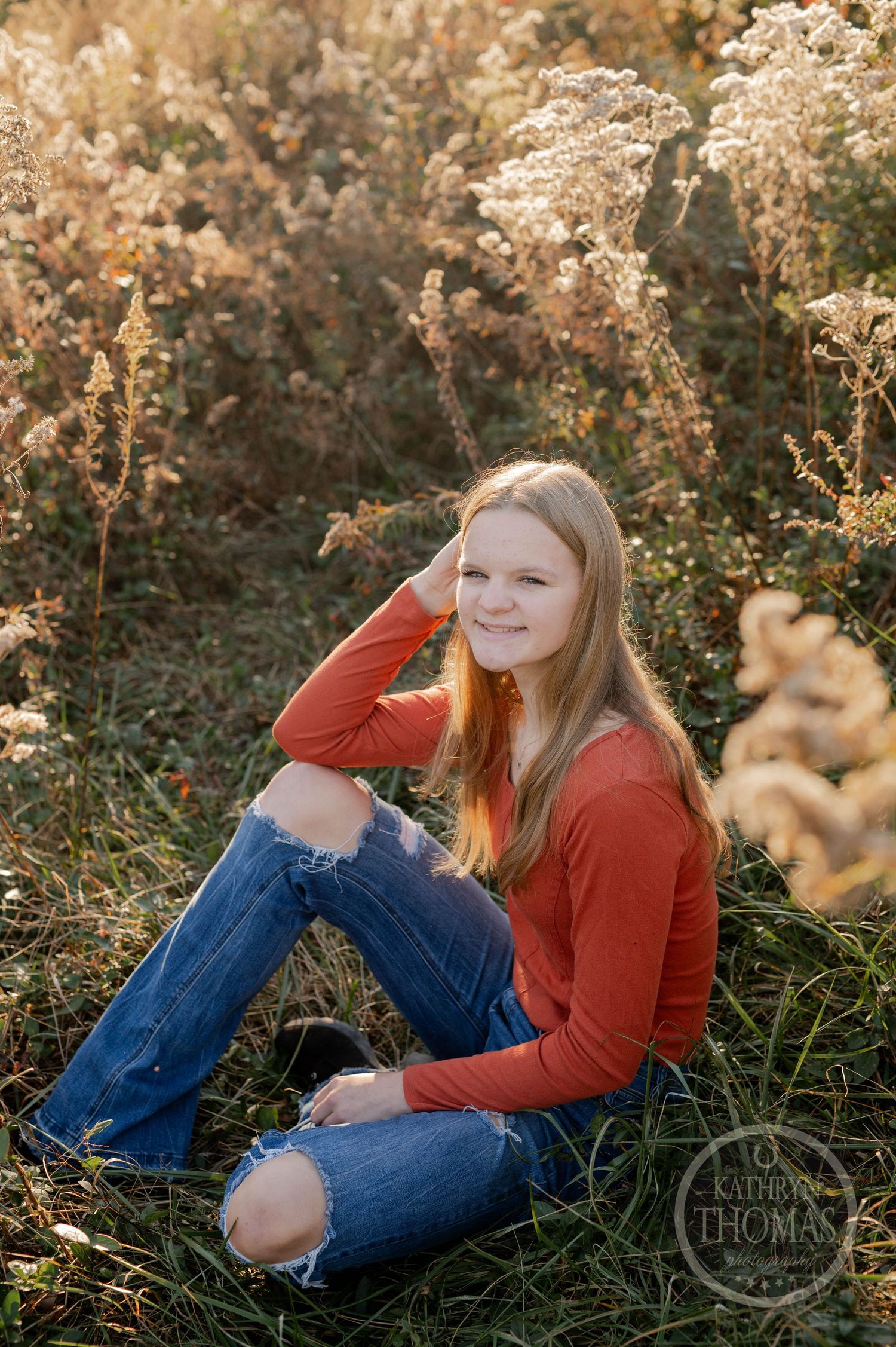 Portrait of someone in an orange sweater sitting in tall autumn grasses at golden hour.