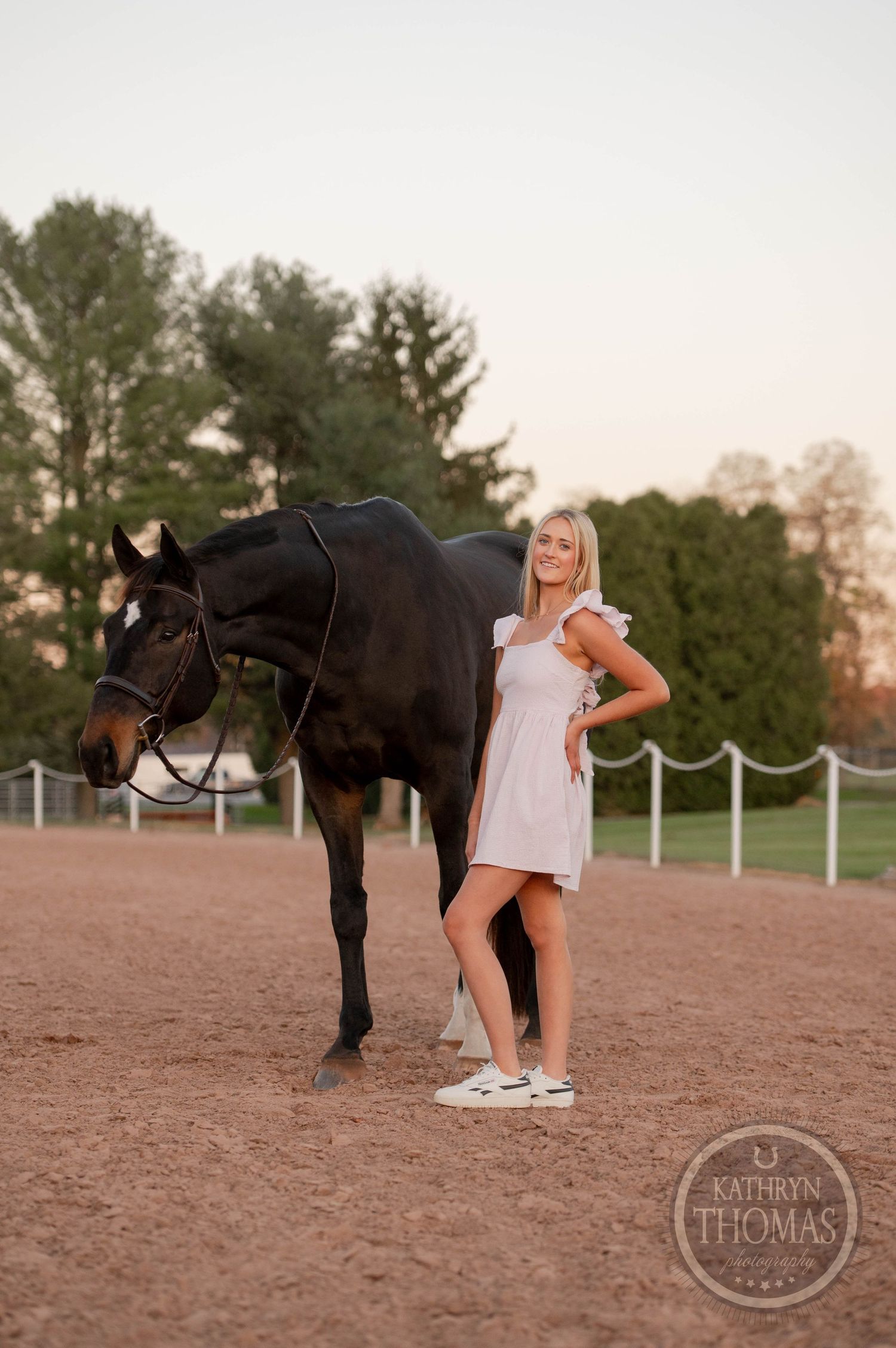 A person in a white dress poses with a black horse in an outdoor riding arena.