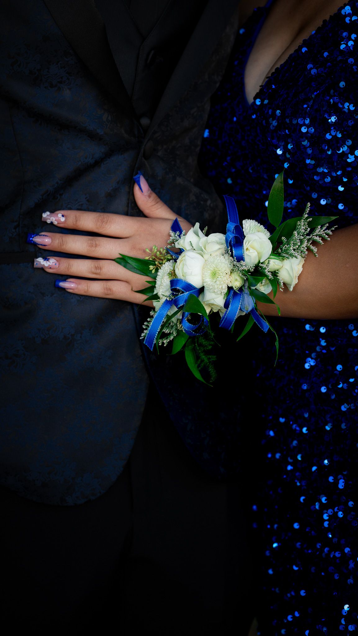 A wrist corsage with white roses and blue accents rests on a hand wearing navy blue nail polish and formal attire.