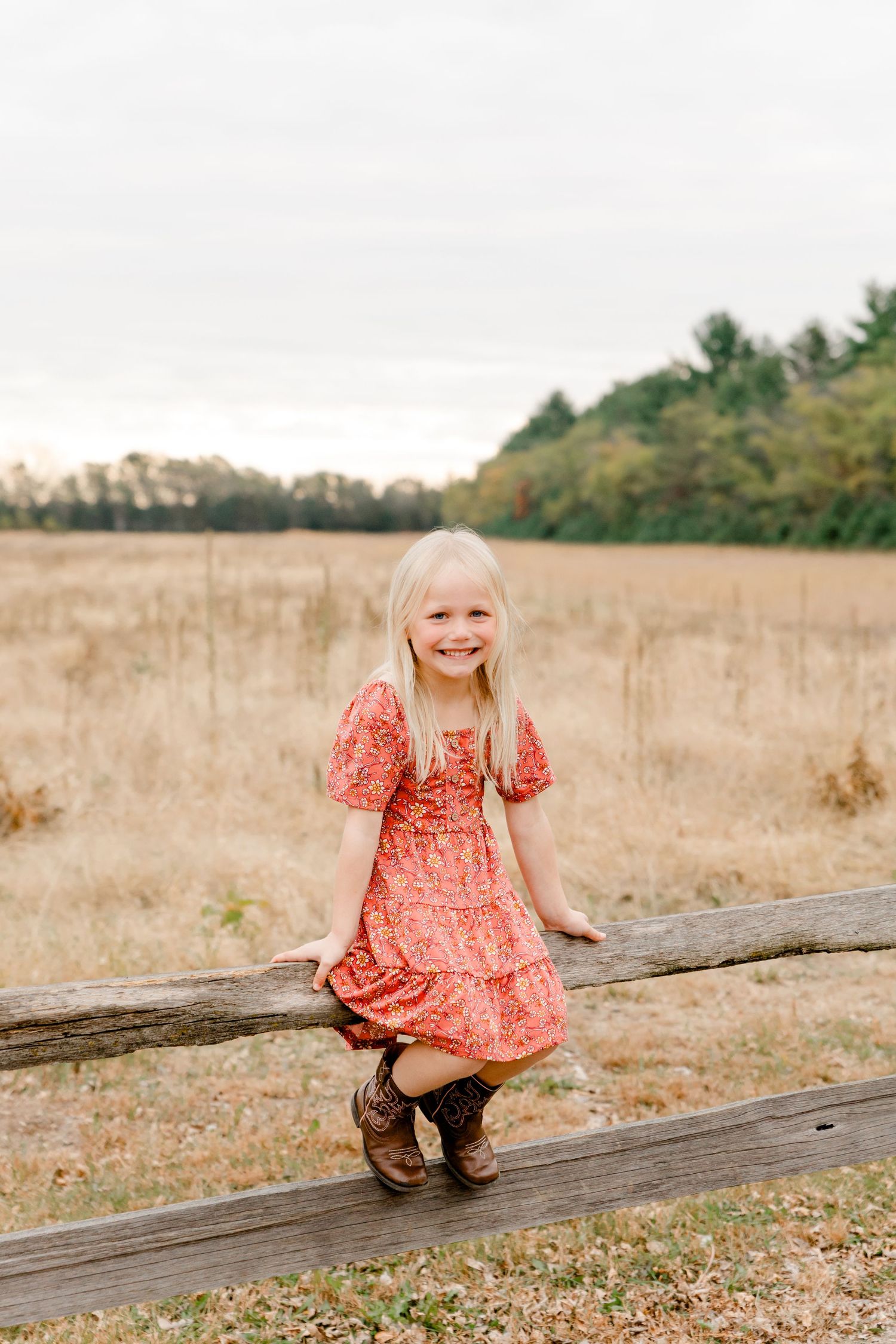 Smiling blonde girl in red floral dress sitting on wooden fence rail in autumn field.