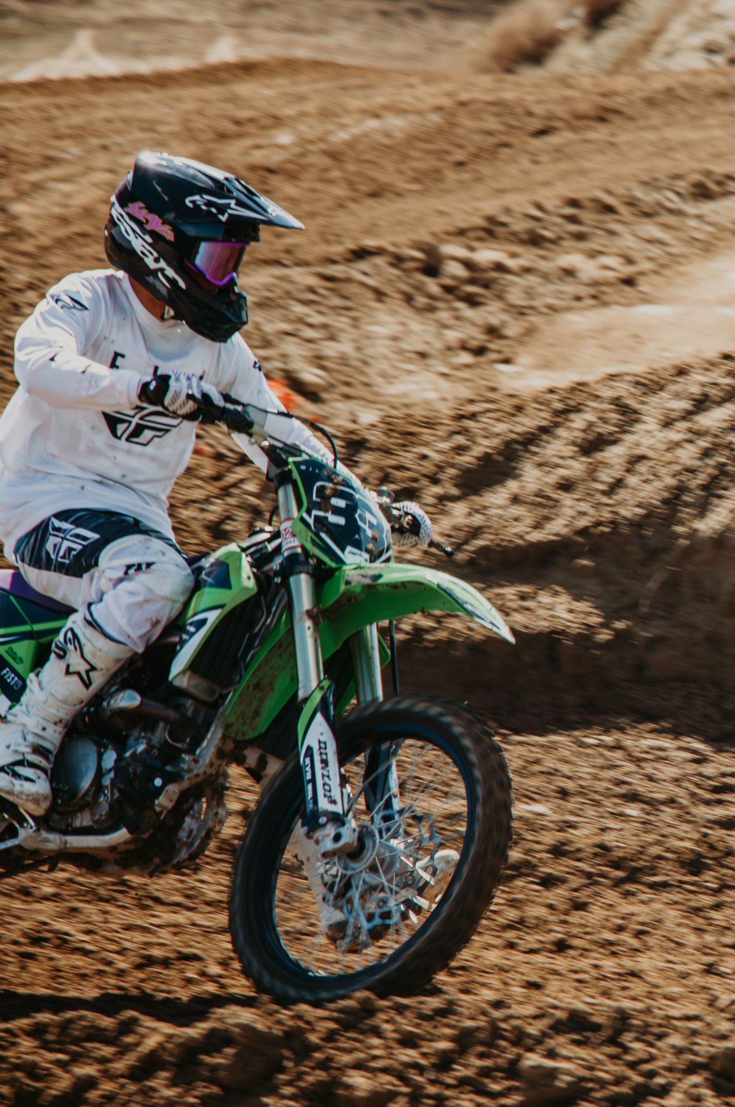 A motocross rider in white gear navigates a dirt track on a green Kawasaki dirt bike during competition.
