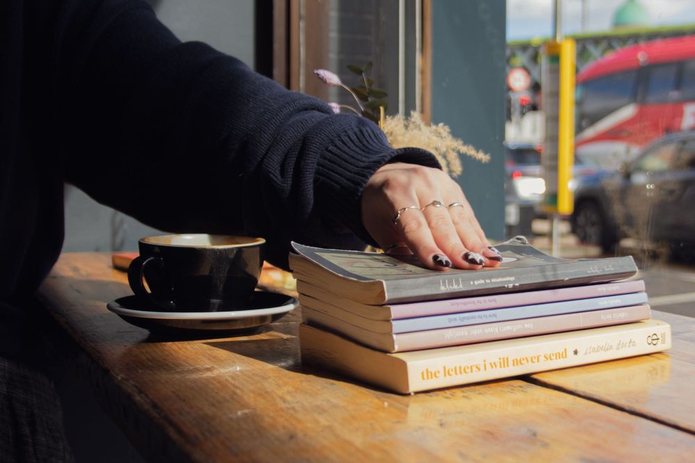 Hand rests on stack of books next to coffee cup on wooden table.