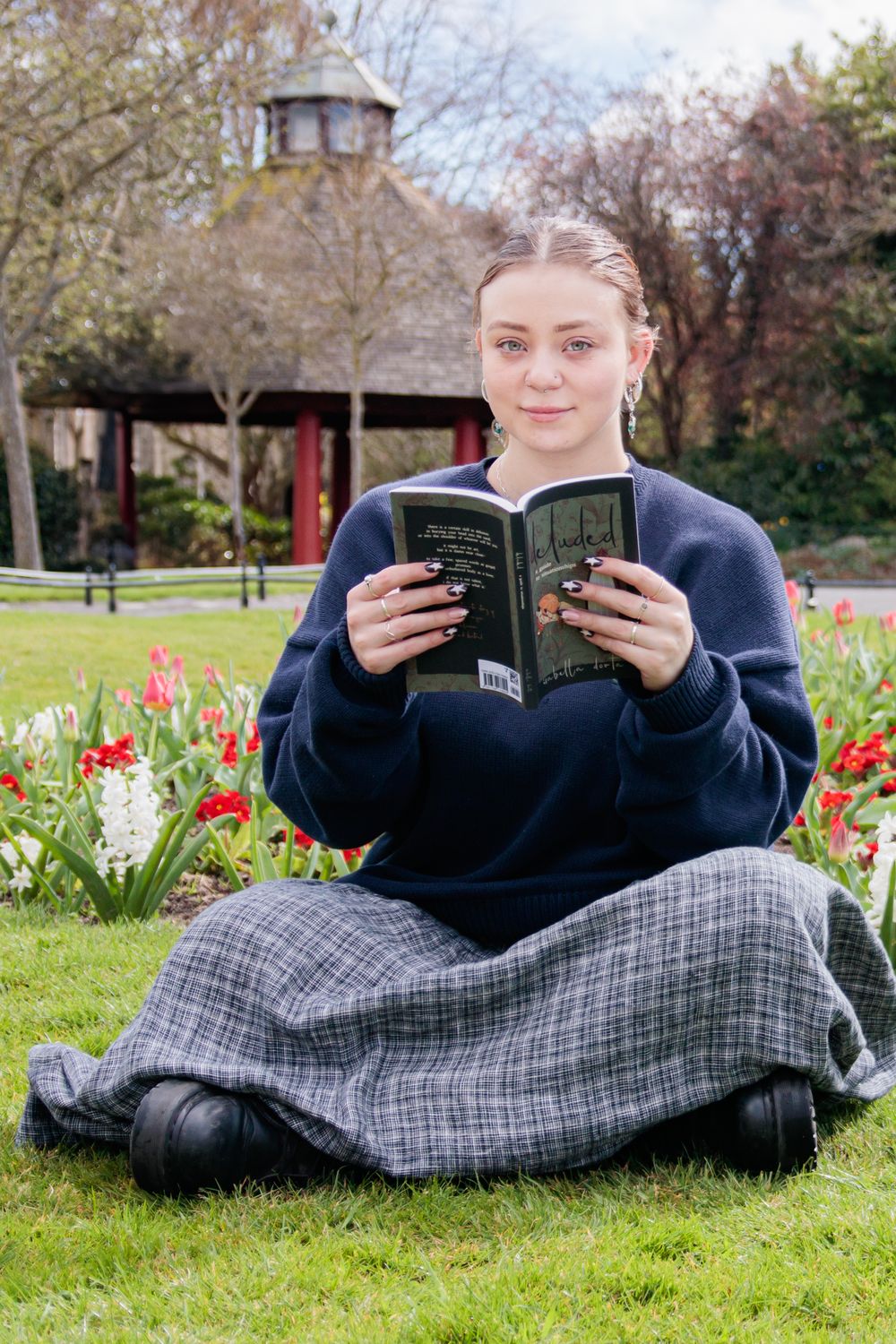 A person reading a book while sitting on grass with tulips and a gazebo in the background.