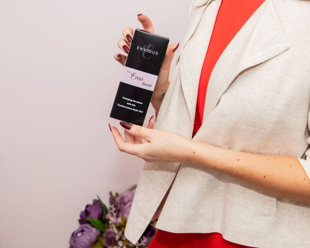 Woman in white blazer and red dress holding small black product box against pink background.