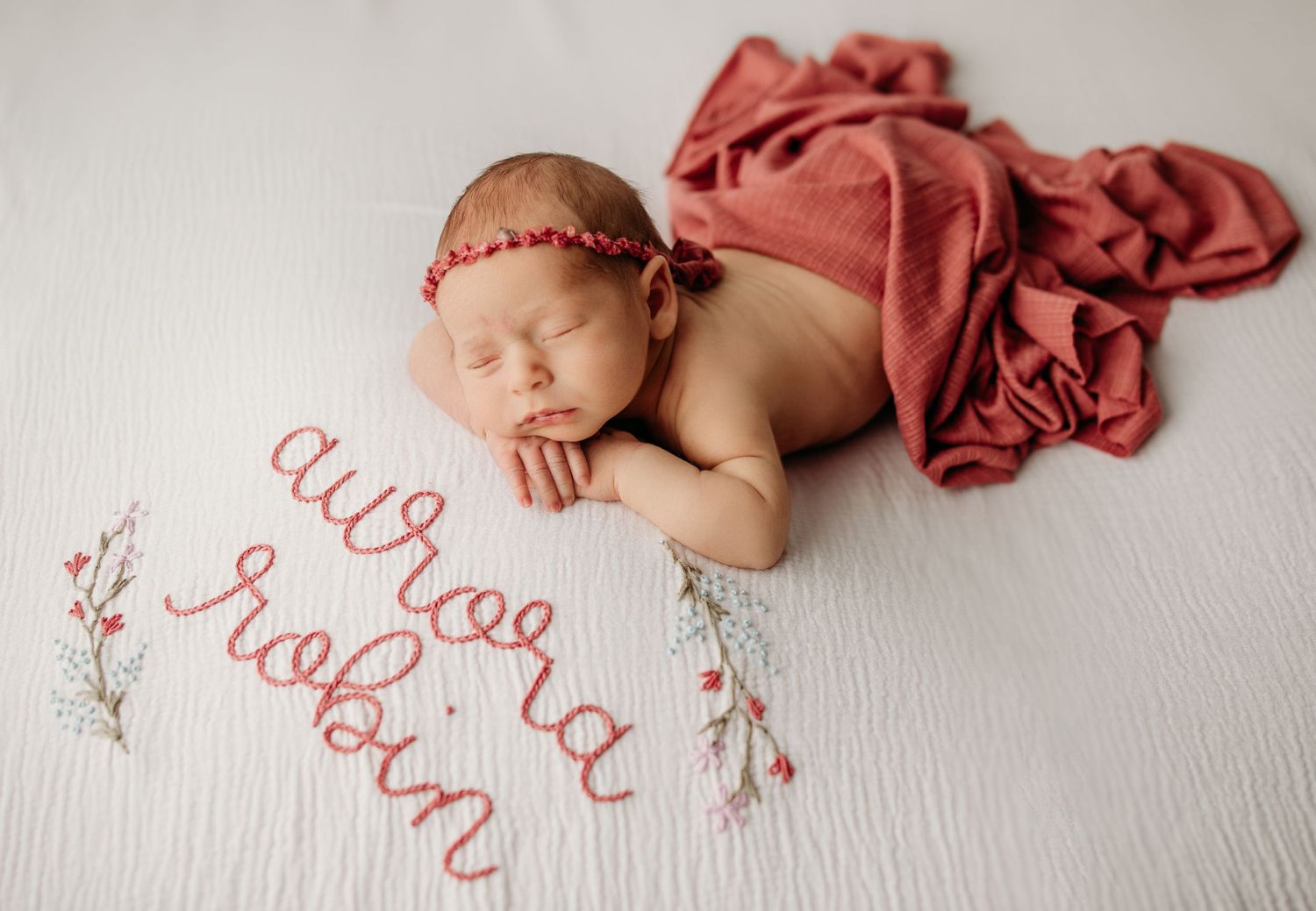 Artistic newborn portrait with coral fabric and embroidered text details on white background.