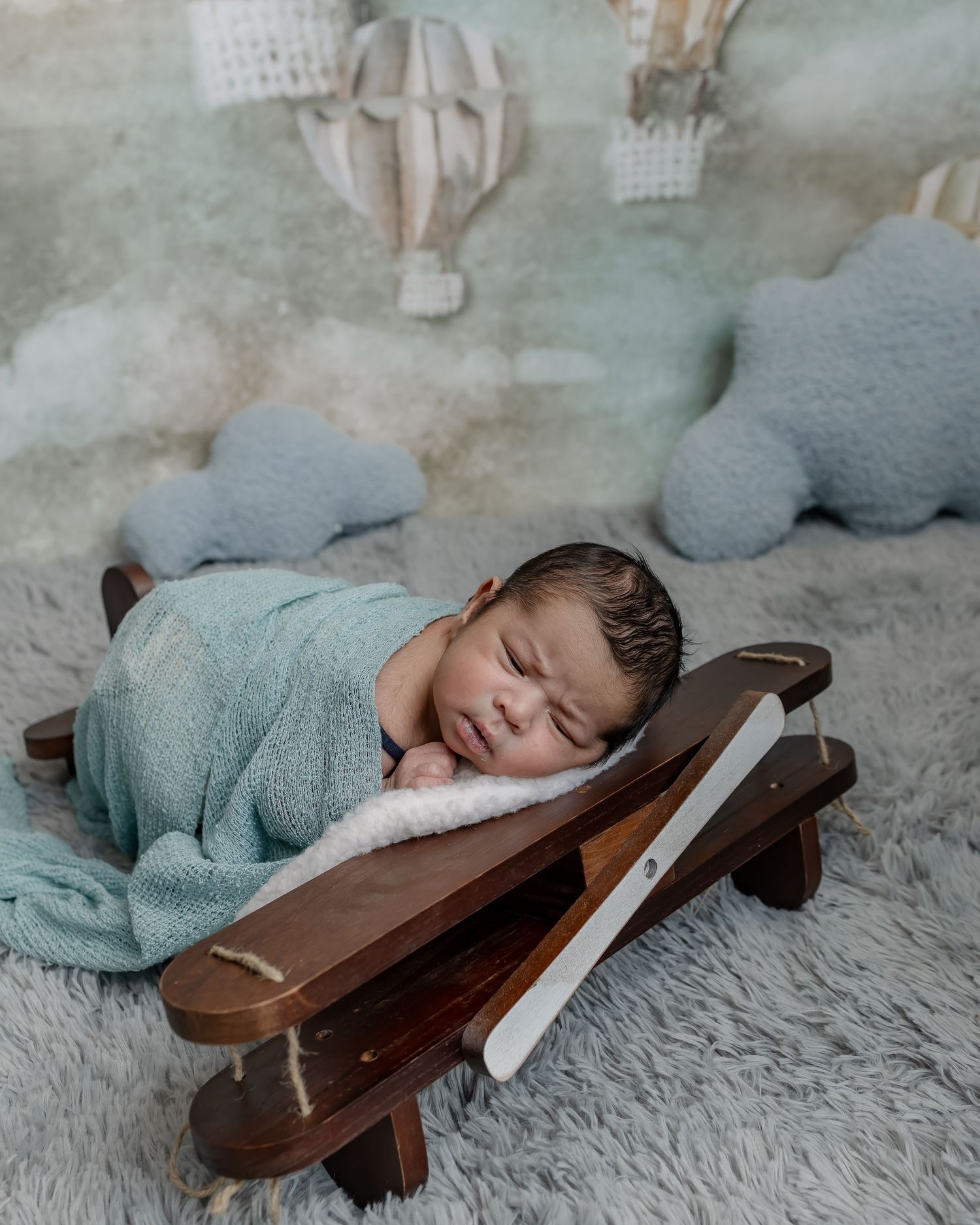 Sleeping newborn wrapped in teal blanket lying on decorative wooden airplane prop with hot air balloon decor.