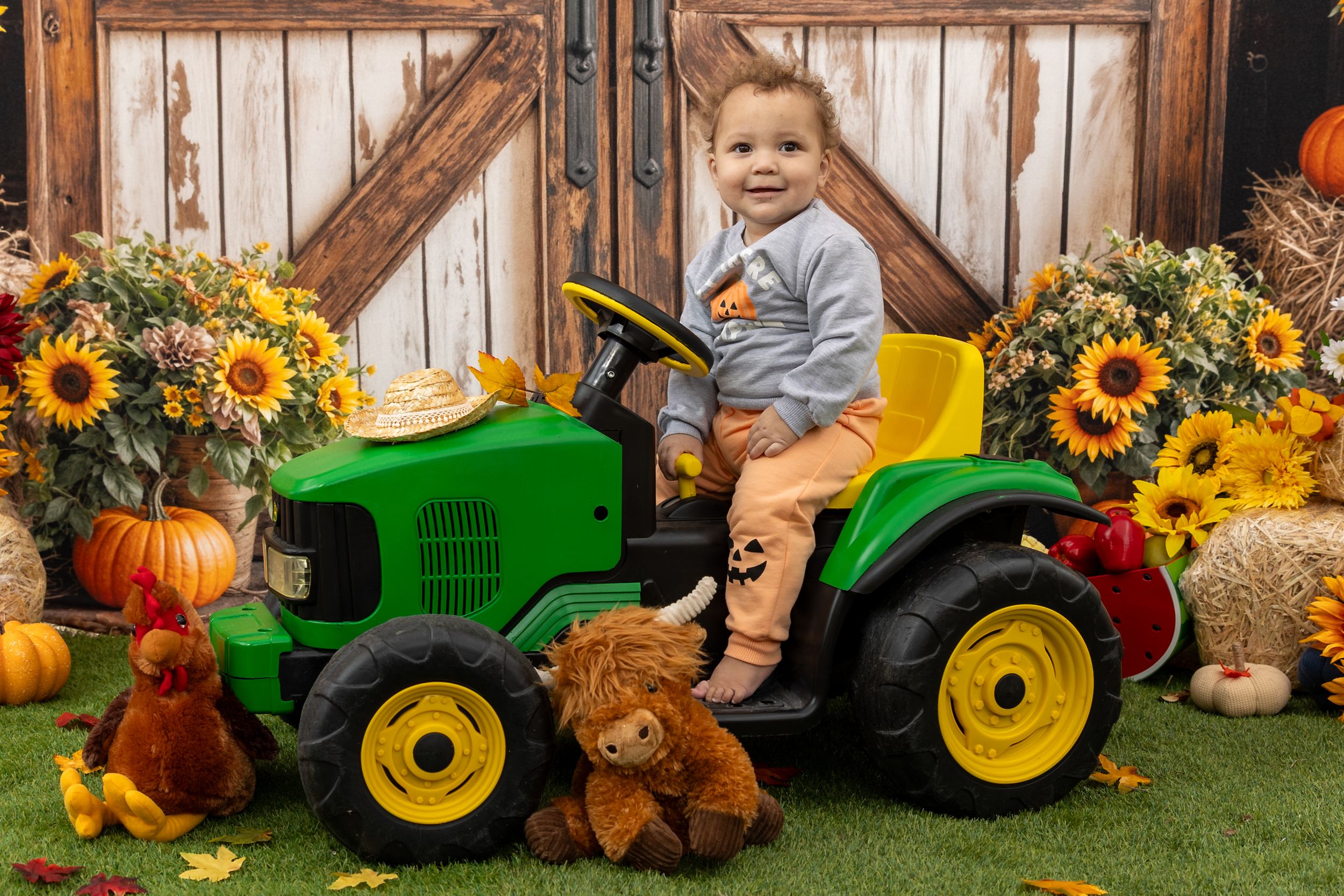 Fall harvest display with green tractor, pumpkins, teddy bears, sunflowers and autumn decor against wooden fence backdrop.