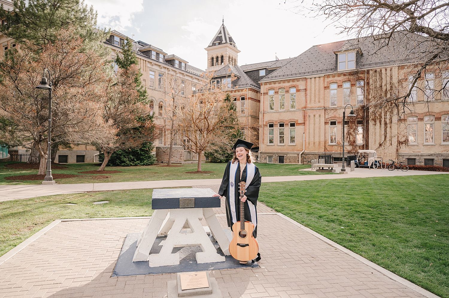 Graduate poses with guitar on college campus walkway with historic brick buildings and bell tower in background.