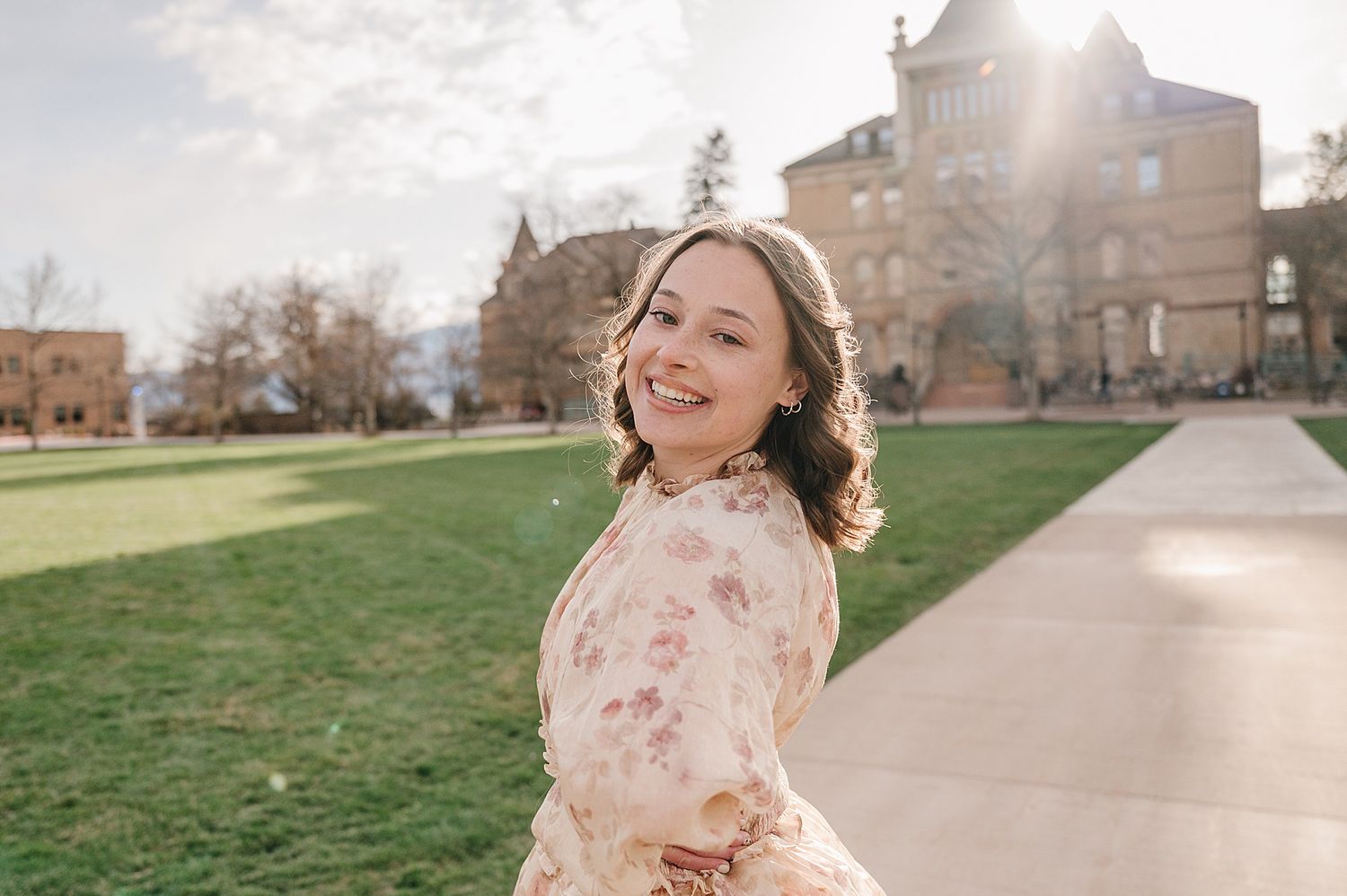 A smiling student in a pink floral dress spins joyfully on a walkway at a university campus on a sunny day.