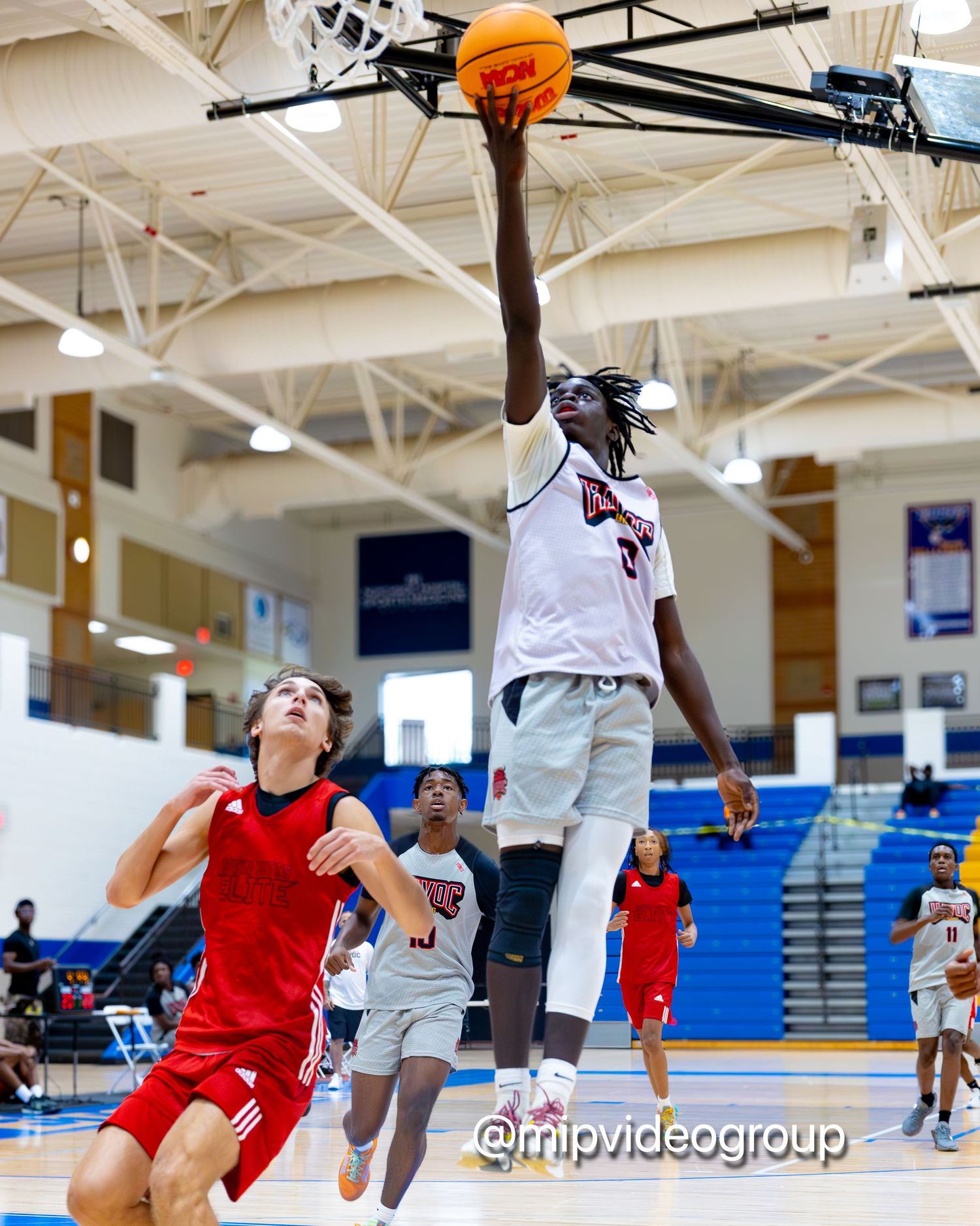 Basketball player in white jersey leaps high for a layup during indoor game against defender in red uniform.