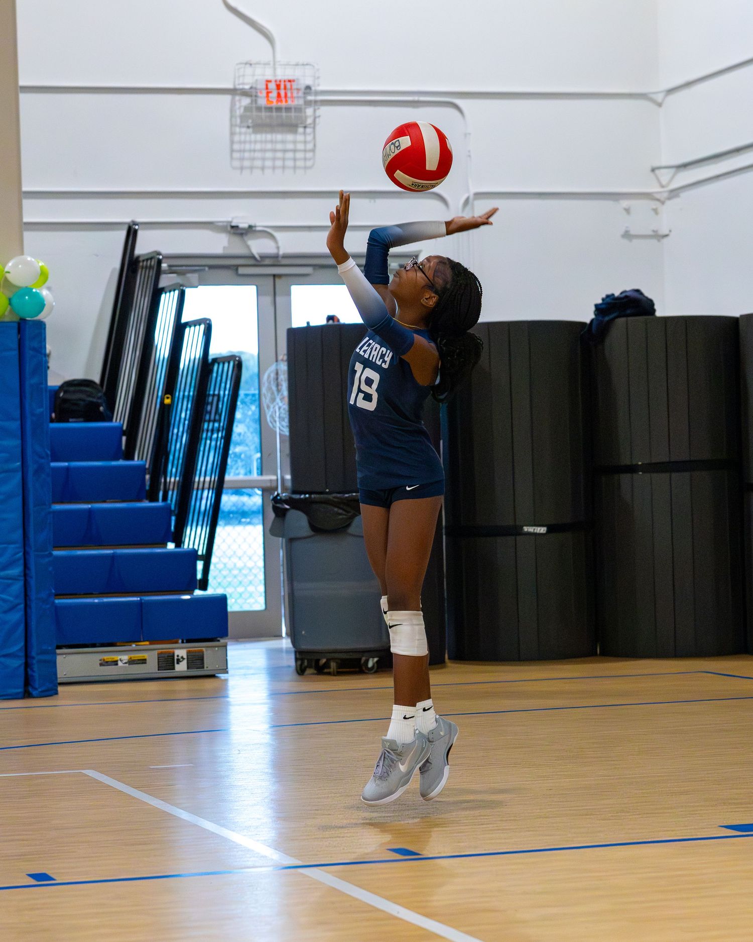 A volleyball player in a navy blue uniform jumps to serve the ball in an indoor gymnasium.