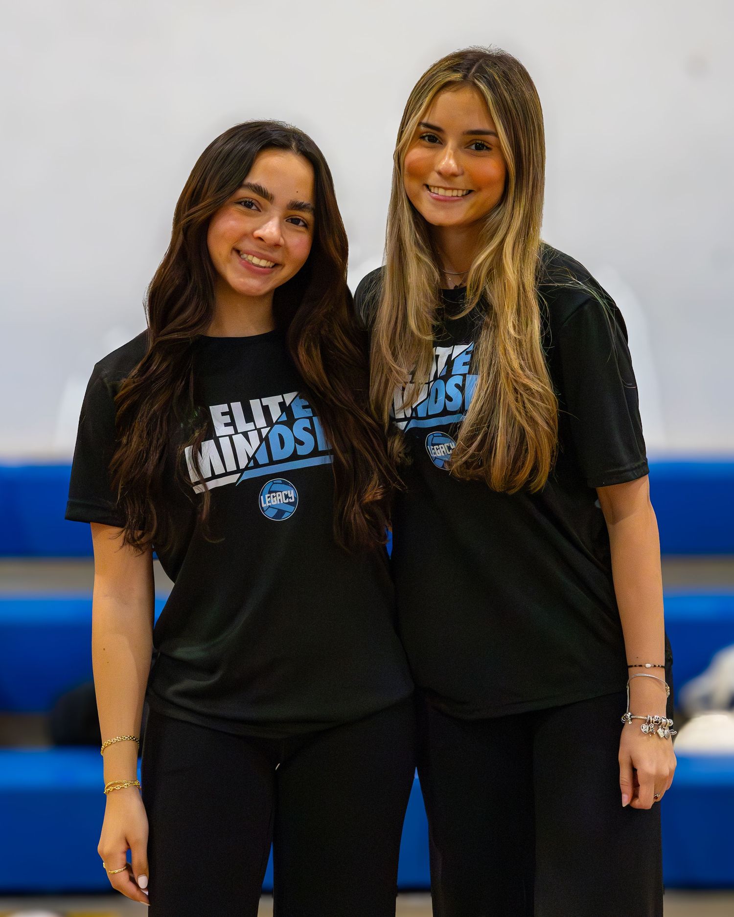 Two people wearing matching black Elite Minds t-shirts pose together in front of blue bleachers.