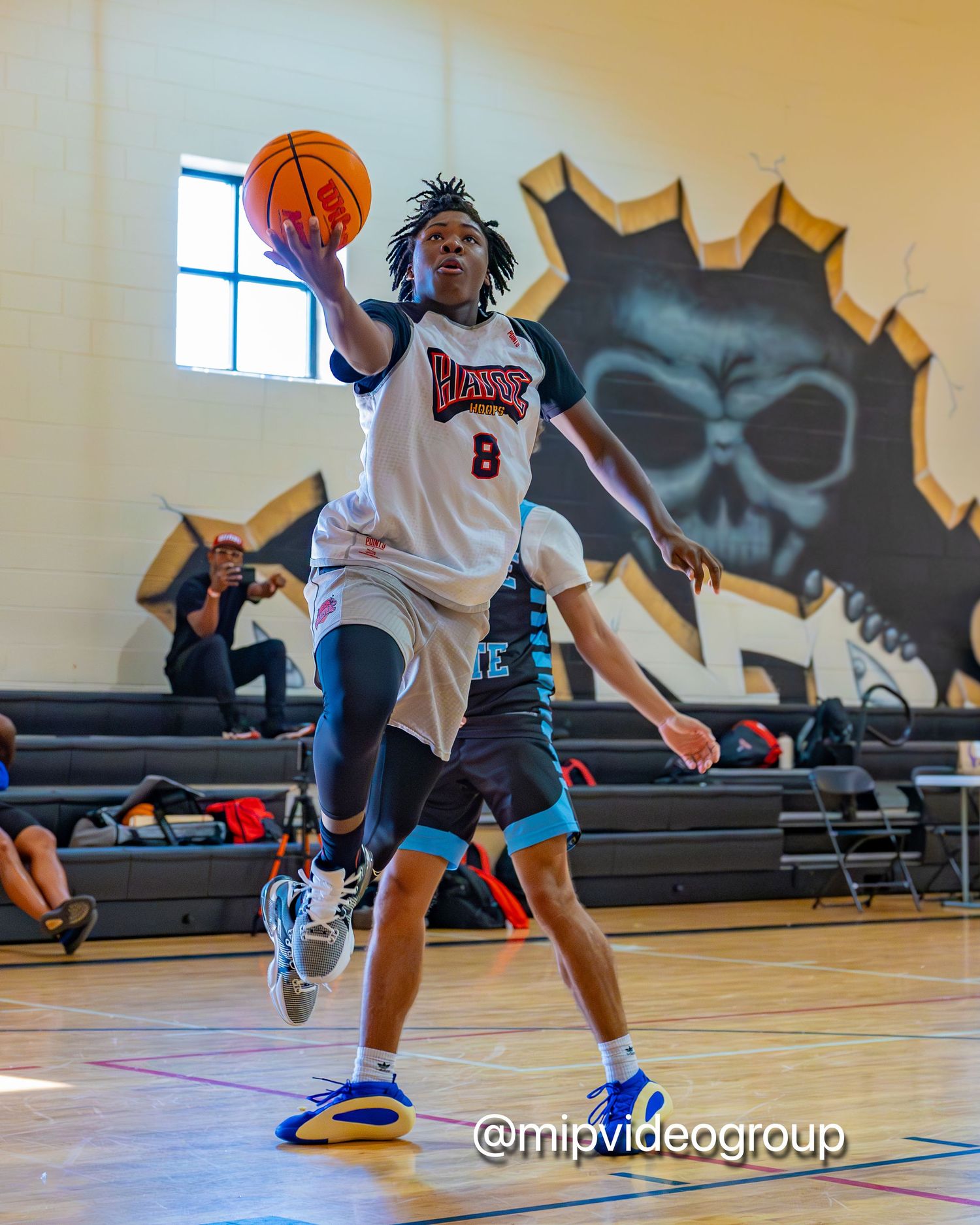 Basketball player in white jersey number 8 leaps for a layup during indoor game with skull mural visible in background.