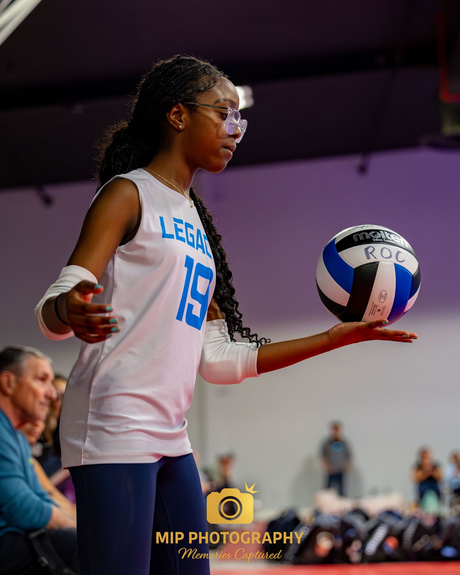 Indoor volleyball player in white jersey and navy shorts jumps to serve the ball during a competitive match.