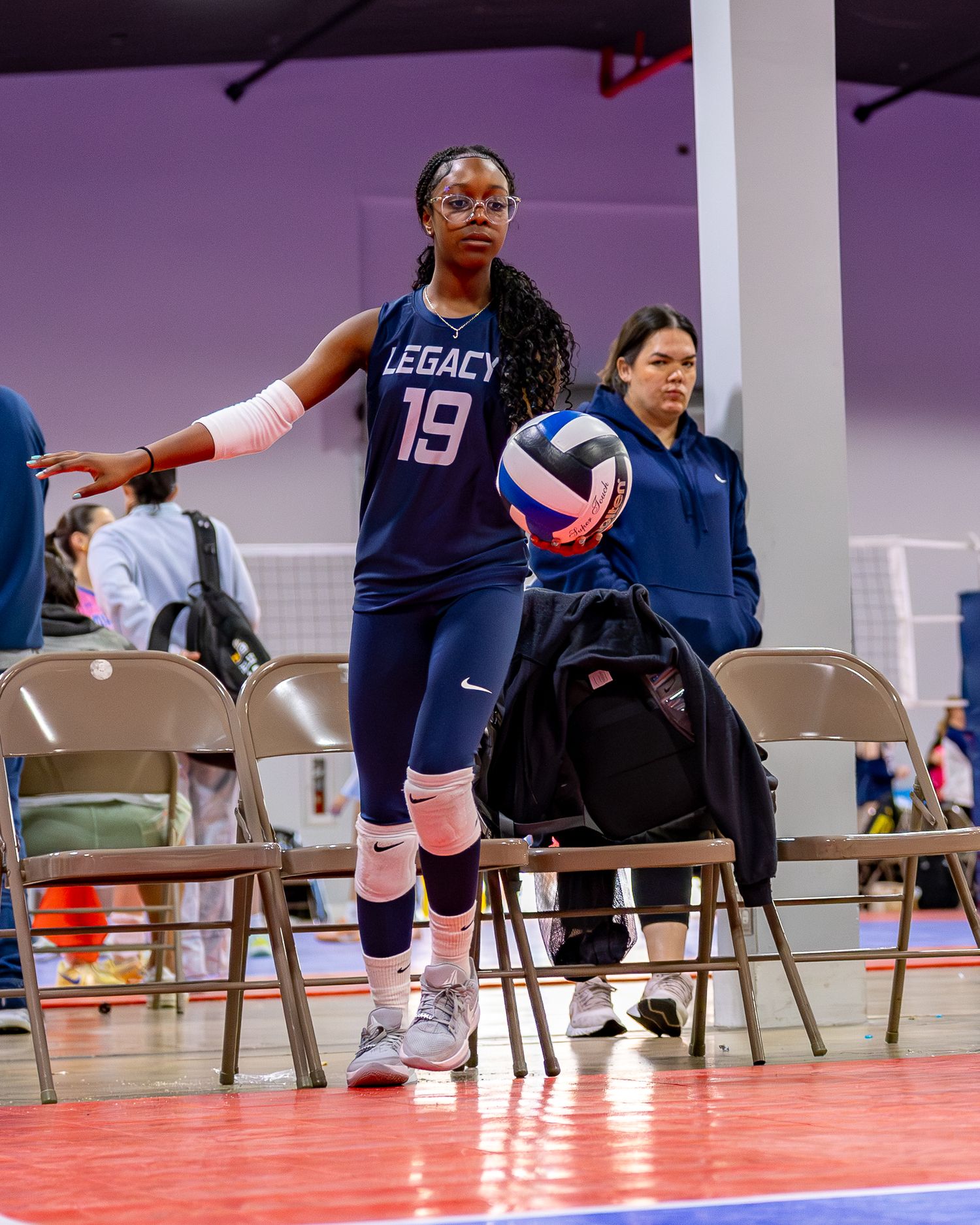 Volleyball player in navy blue Legacy jersey number 19 stands ready with ball during indoor tournament on red court.