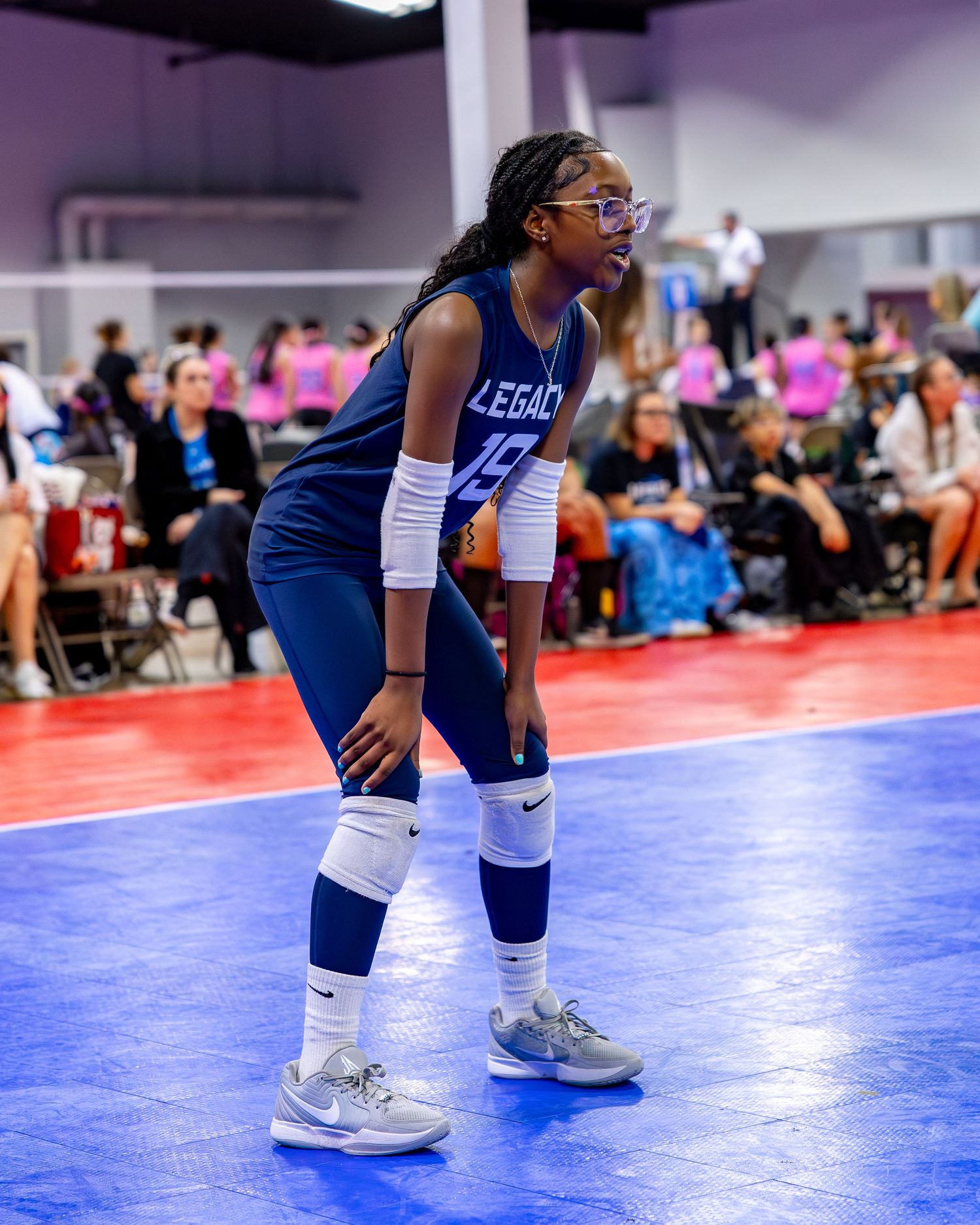 Athletes in navy blue uniforms and knee pads compete on a bright blue volleyball court during an indoor match.