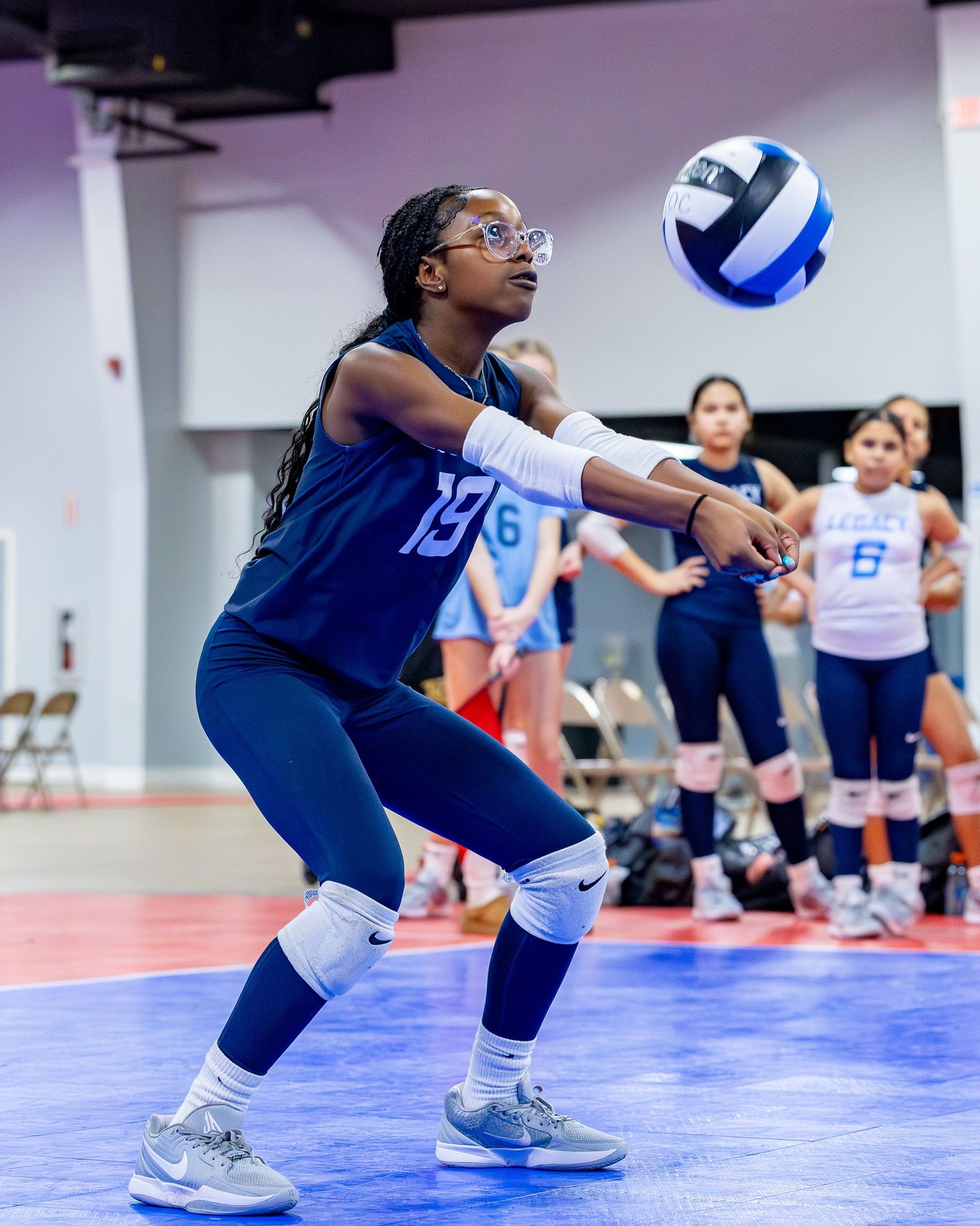 Volleyball player in blue uniform crouches low to receive a serve during an indoor match.