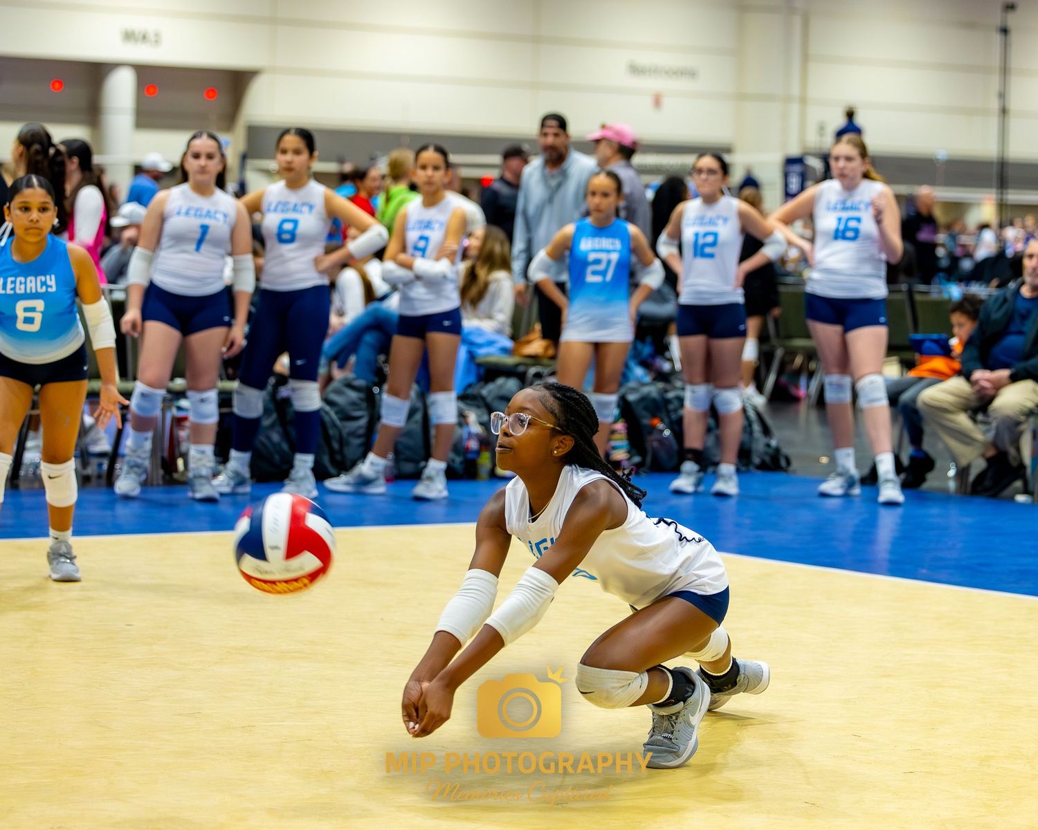 Athletes dive and stretch for a volleyball during an indoor volleyball match on a blue court.