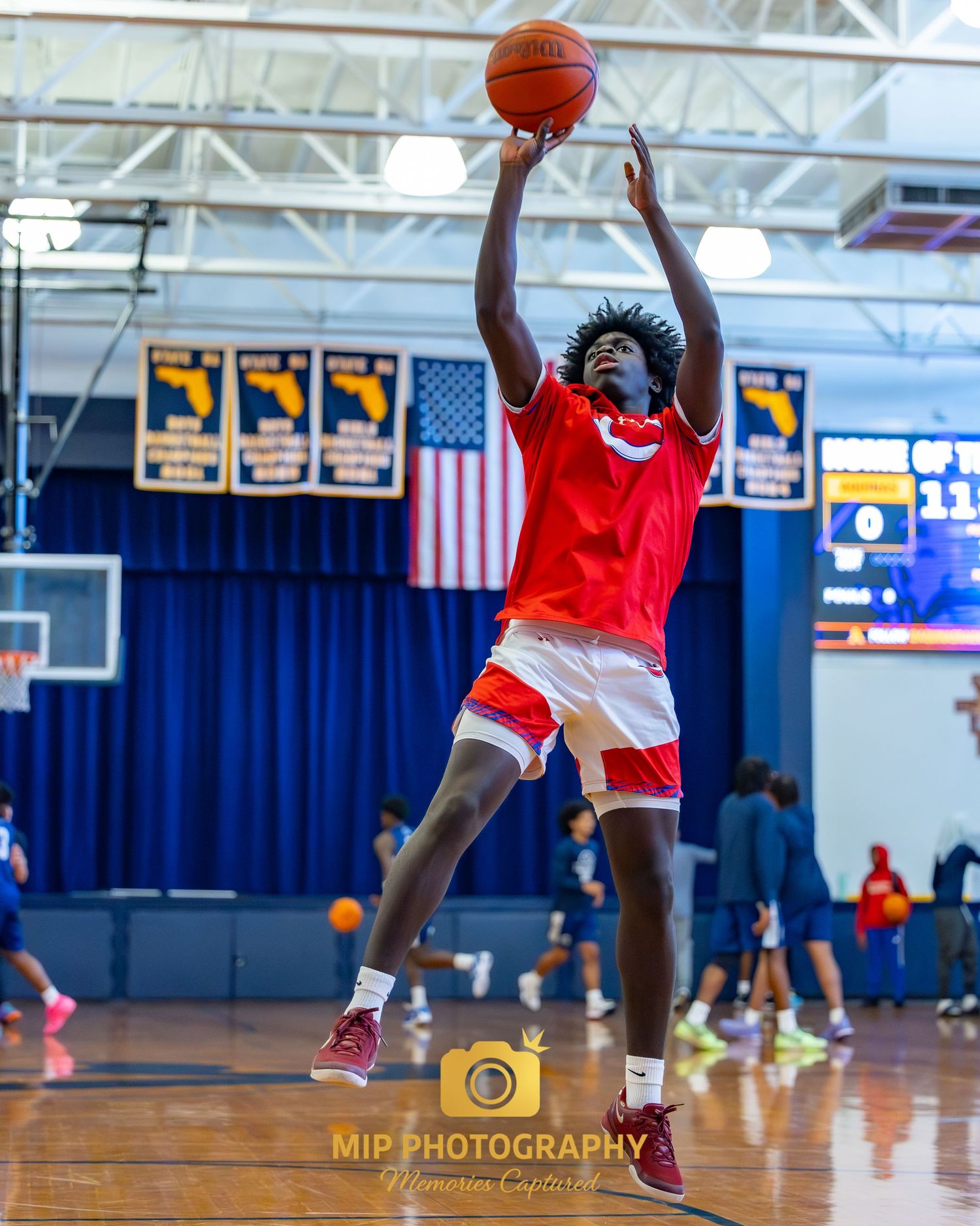 Basketball player in red uniform takes a jump shot in a gymnasium with American flag visible.