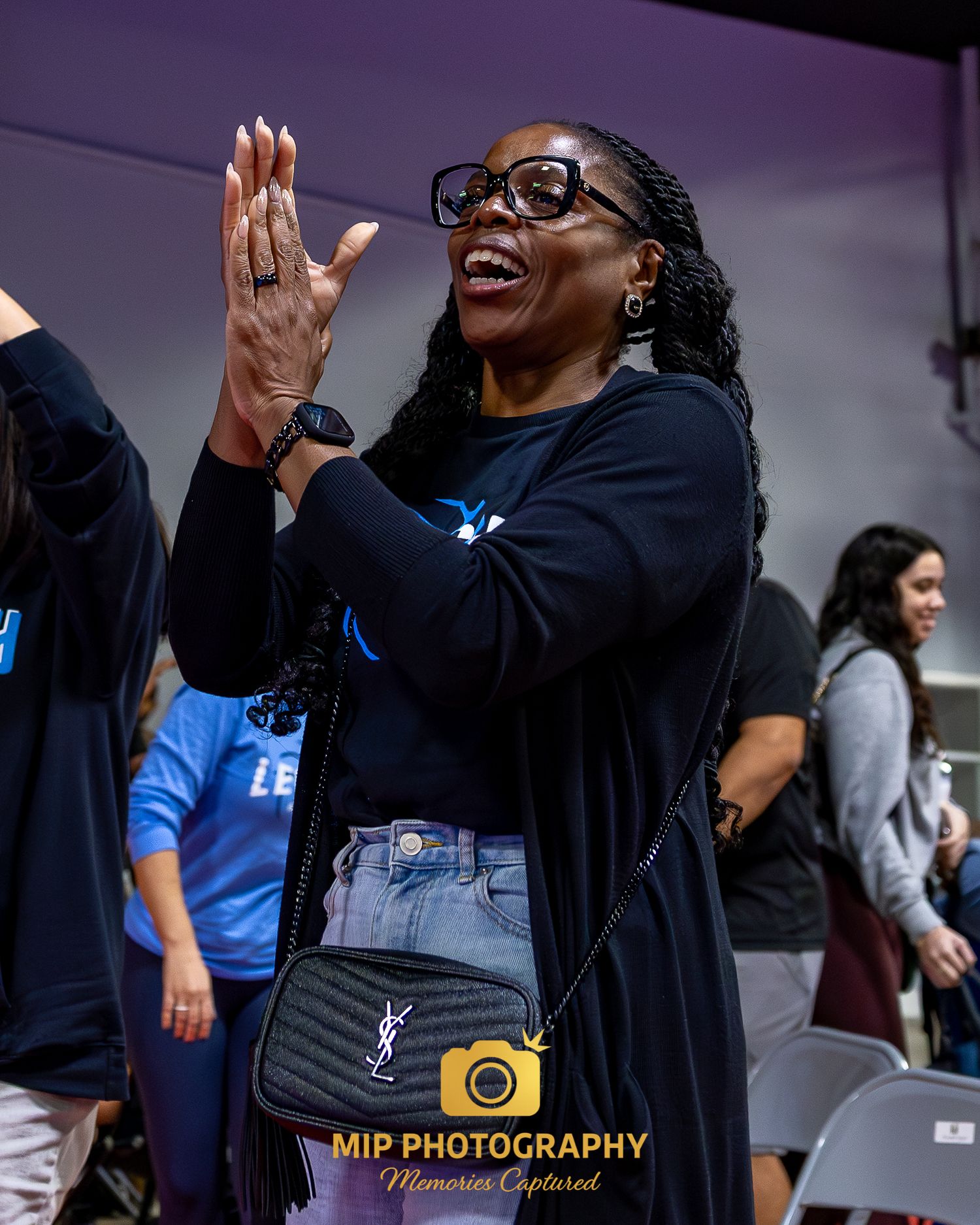 A person in black clothing and glasses gestures enthusiastically while speaking at an outdoor event.