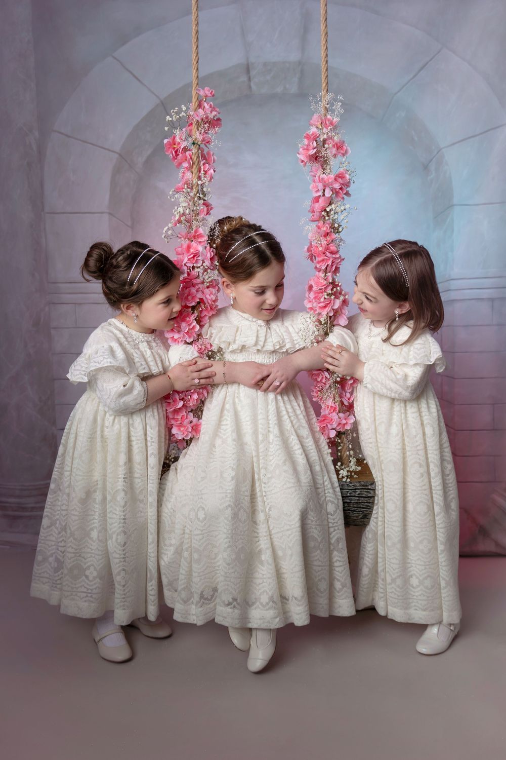 Three children in white vintage dresses pose together by floral swing with pink flowers.