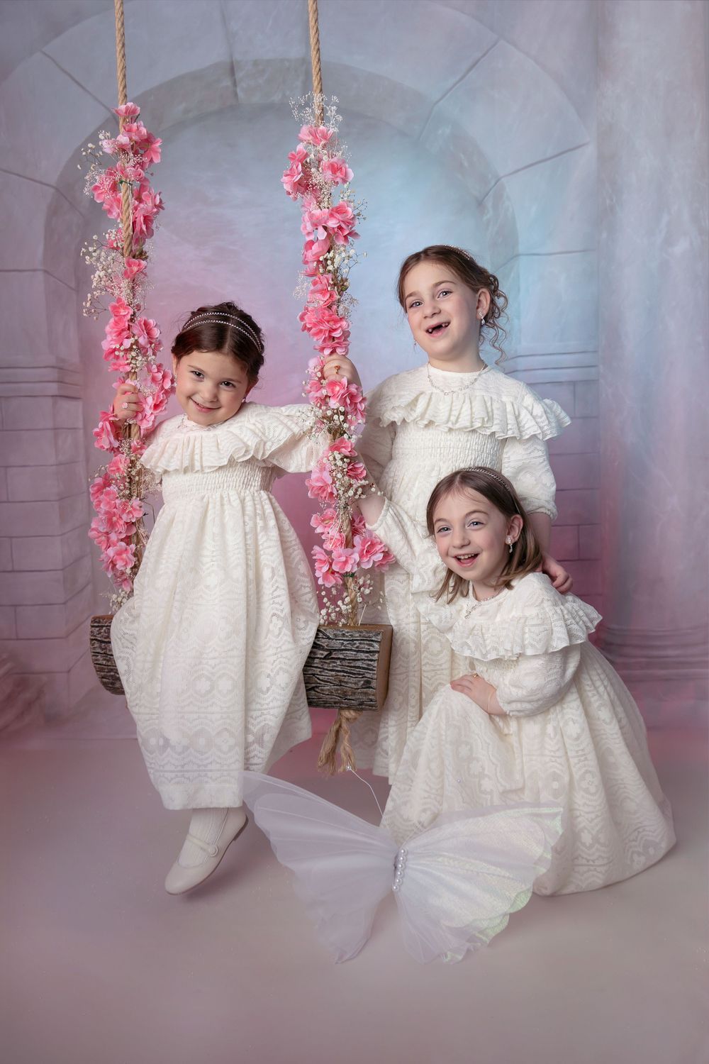 Children in white princess dresses play on fantasy floral swing decorated with pink blossoms in studio setting.