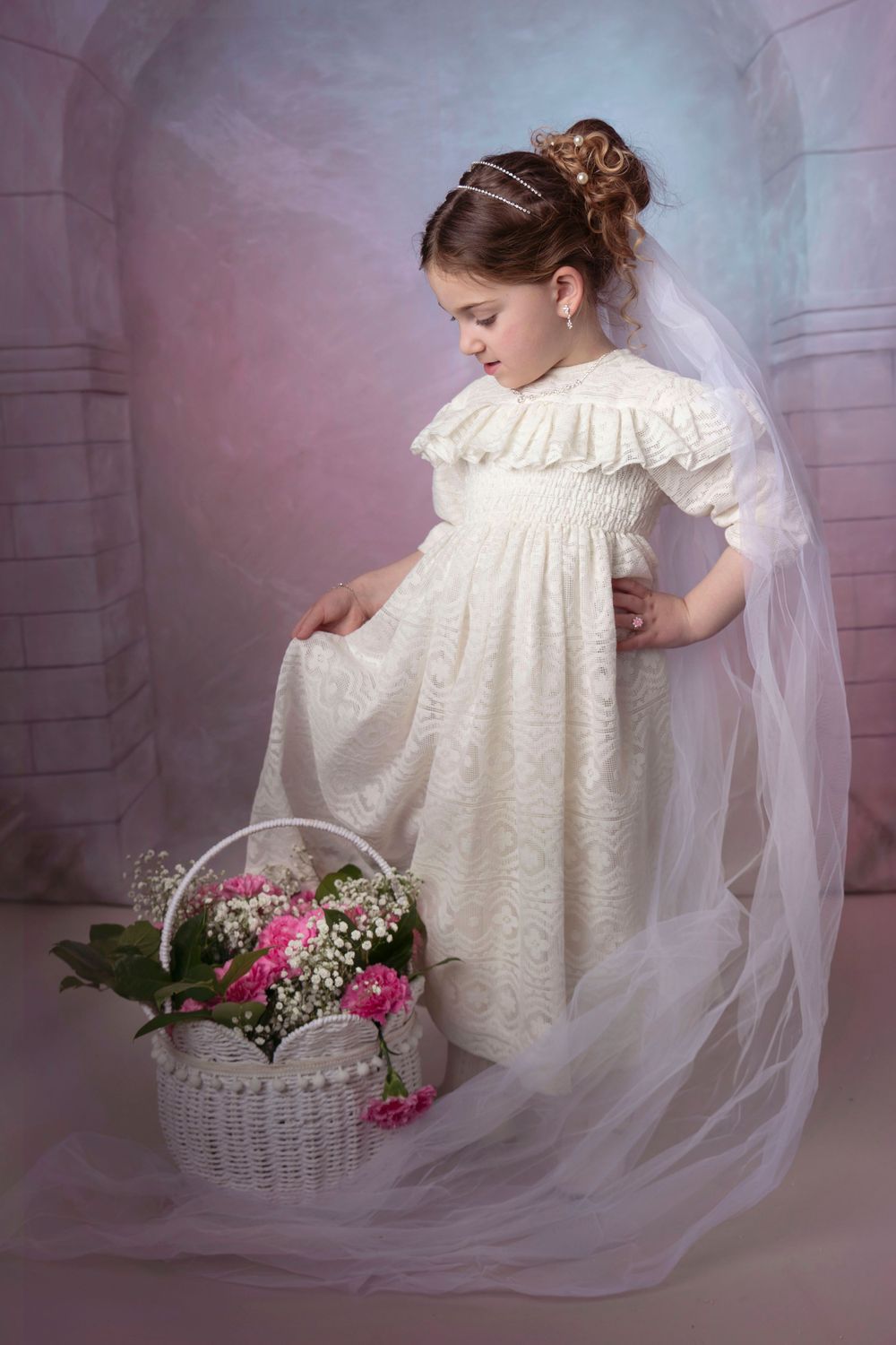 Purim portrait of little girl white lace dress, veil and flower basket against stone arch backdrop.