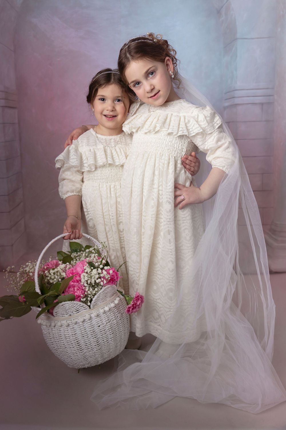Two matching white dresses and flower basket against stone arch backdrop. Purim photo.