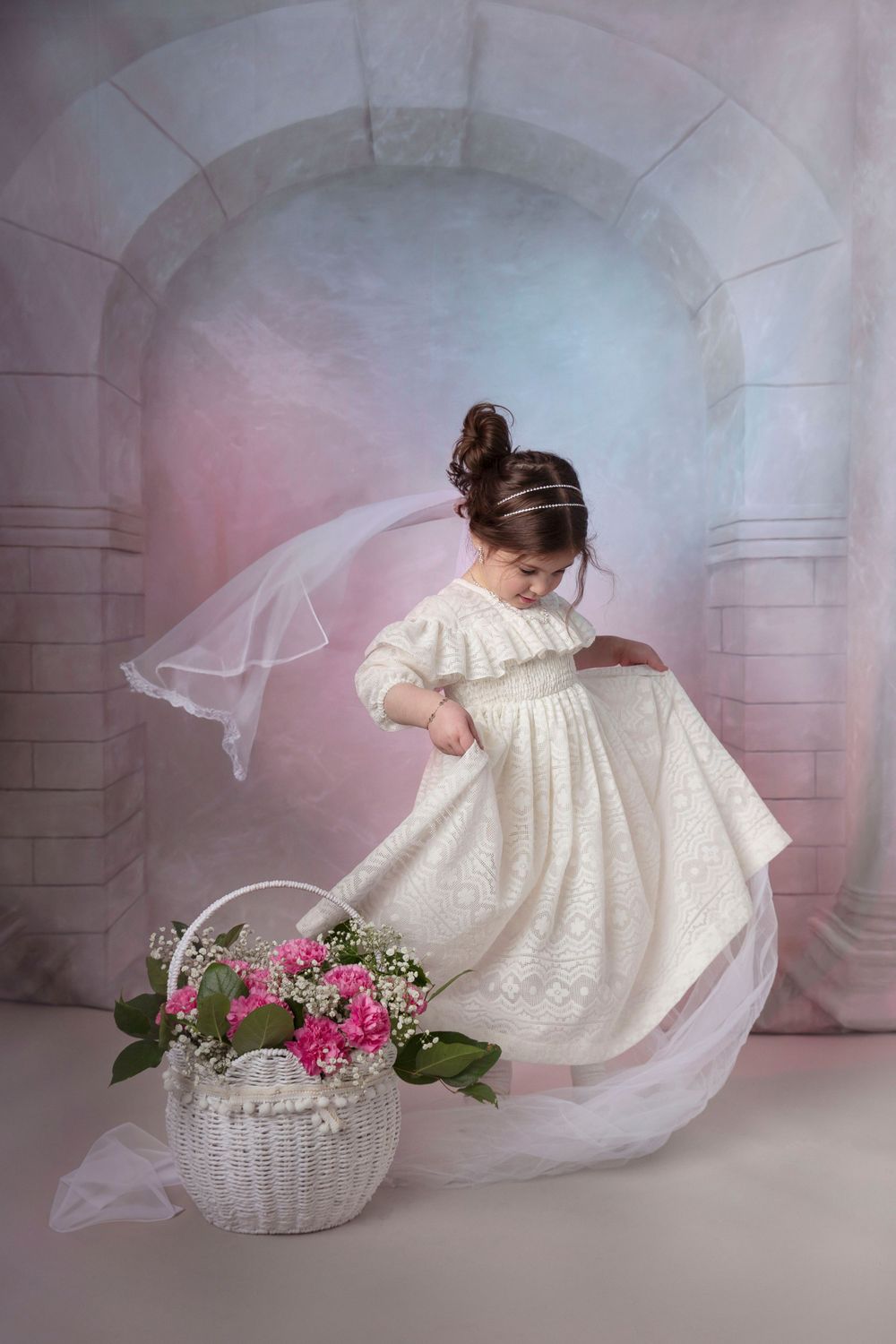 Young girl in vintage white dress and veil twirls next to basket of pink flowers in dreamy archway setting. Purim photo.