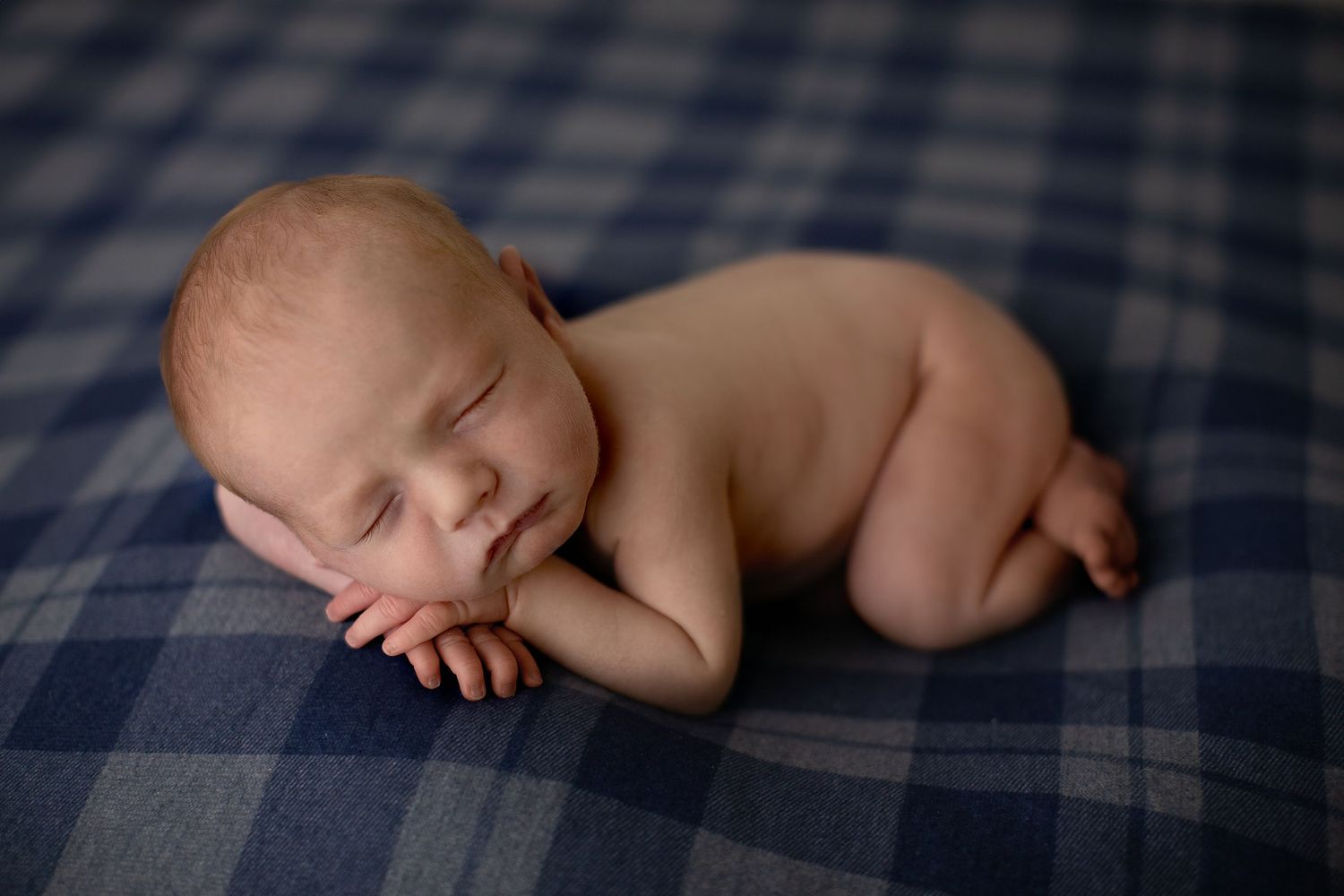Sleeping newborn baby curled up on blue plaid blanket during peaceful professional photography session.
