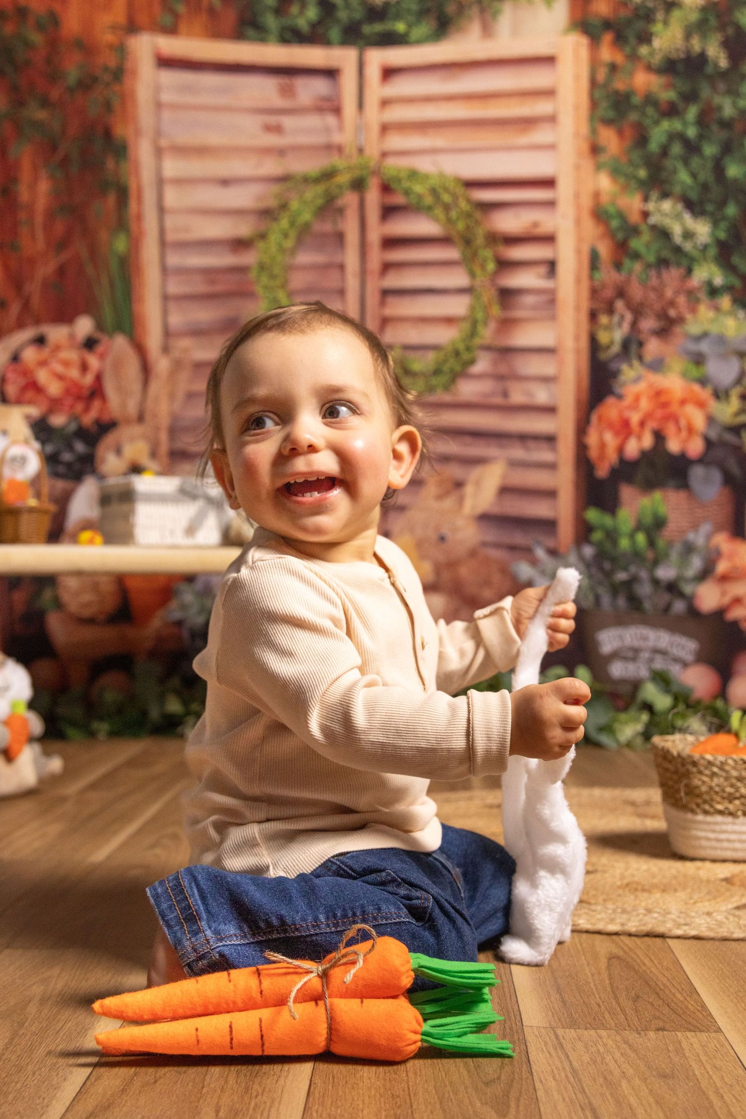 Baby in beige outfit sitting on wooden floor with toy carrots in autumn-themed photo setting.