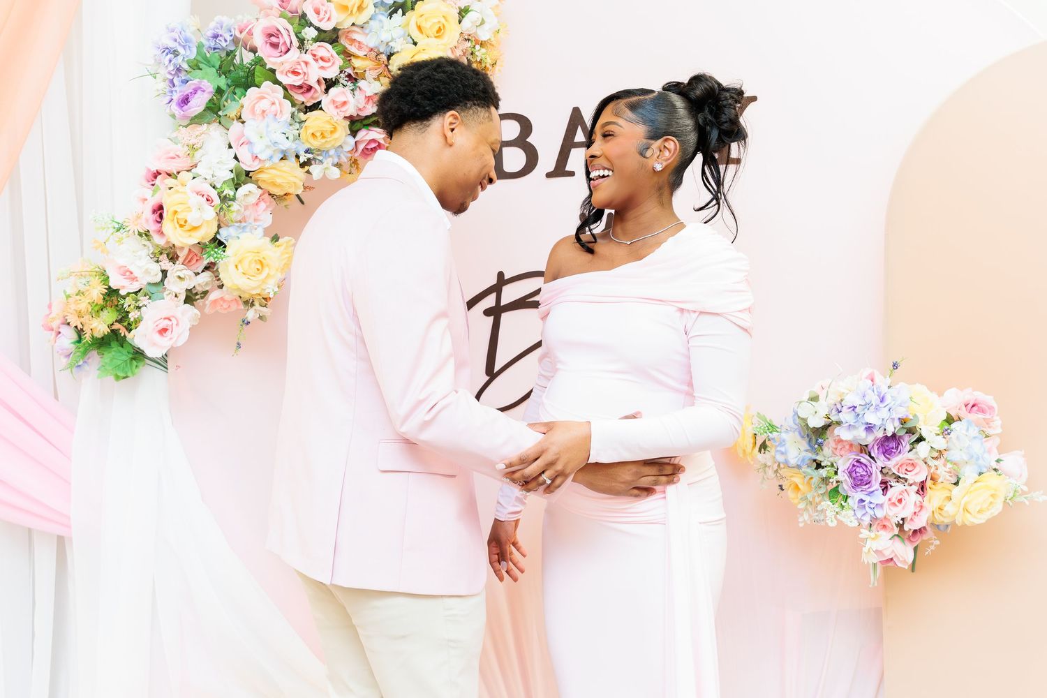 Couple holding hands and smiling at each other at a colorful floral baby shower backdrop.